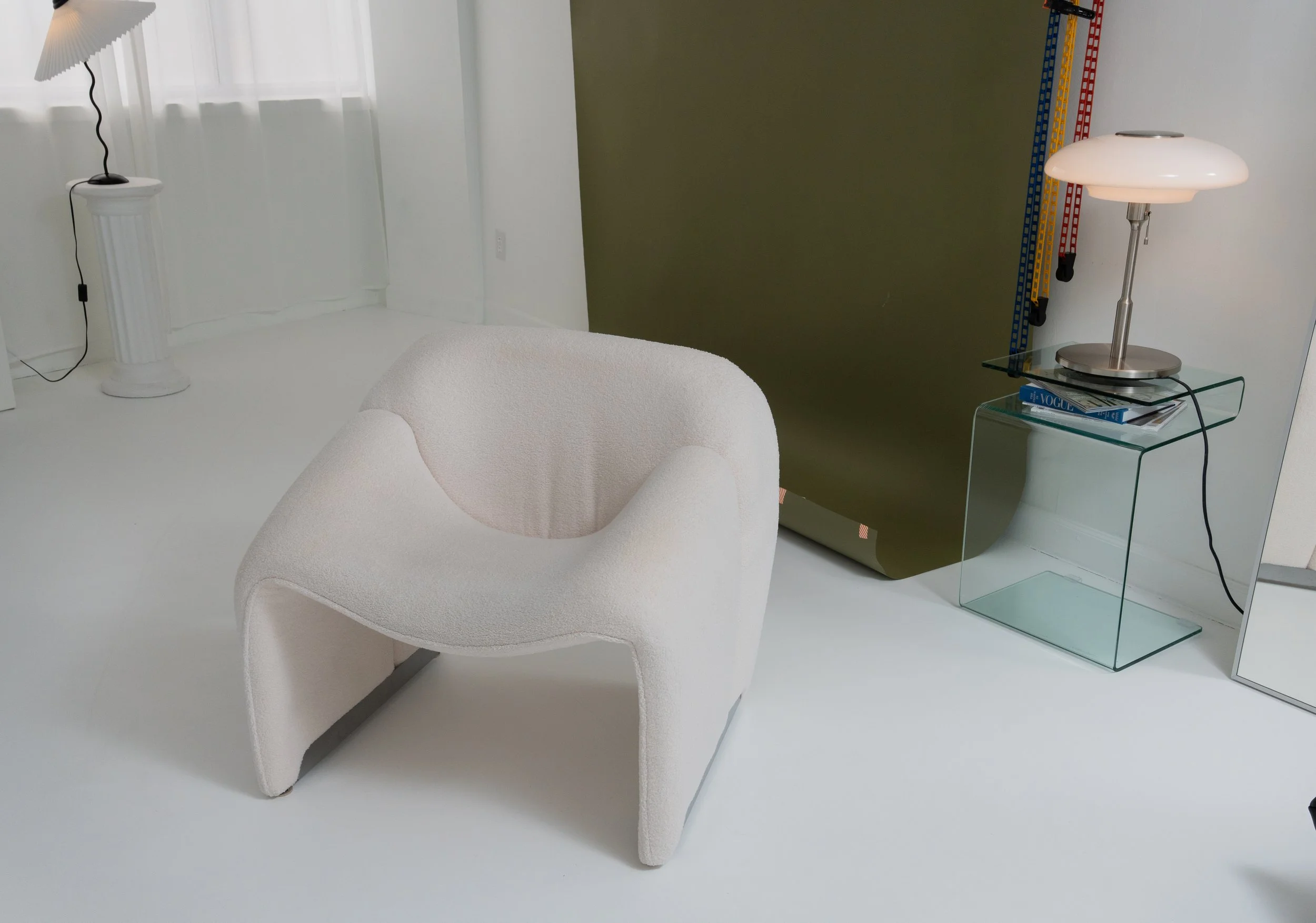 Modern white armchair with a textured fabric, beside a glass side table with a reading lamp and magazines, in a minimalist room with white walls and flooring.