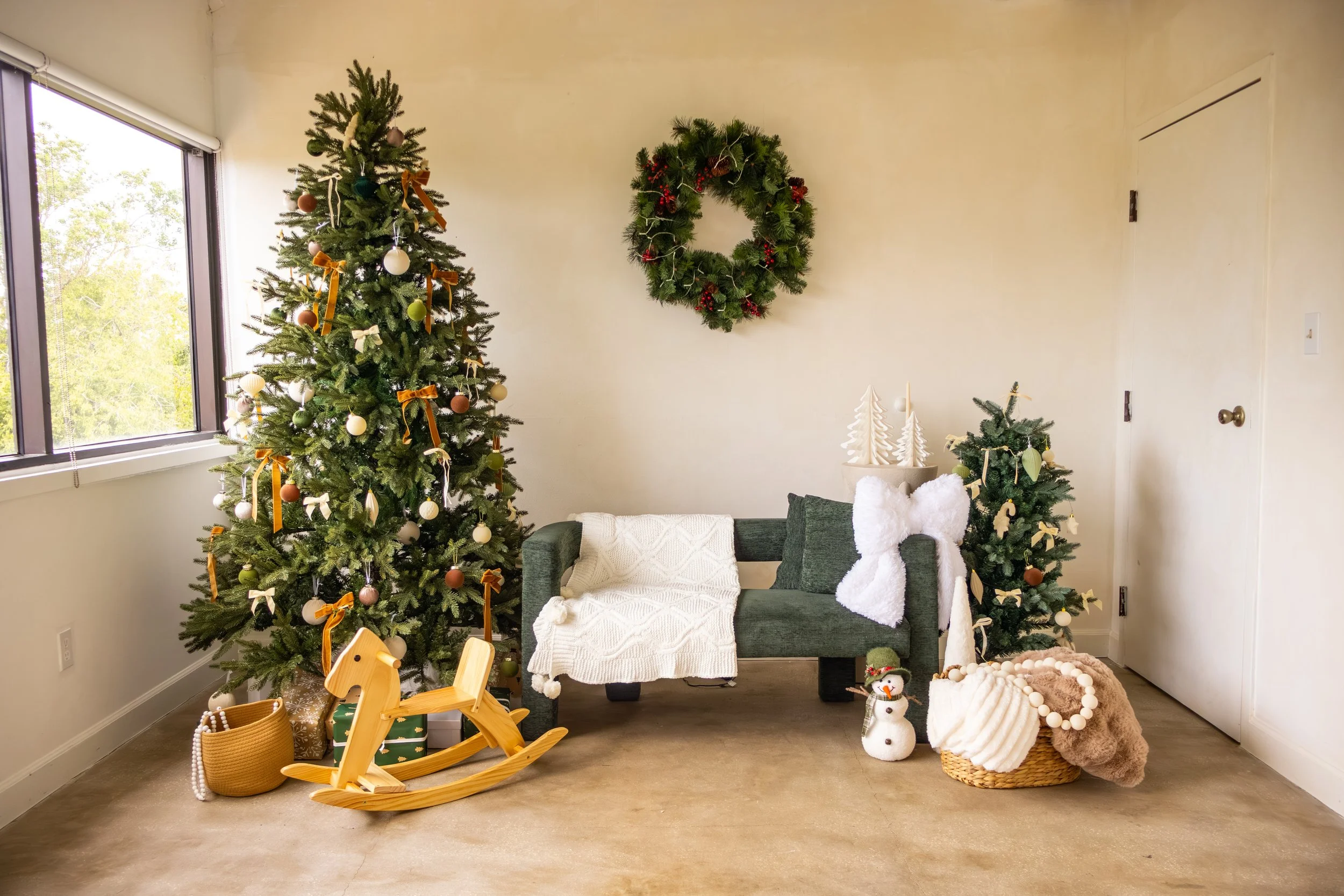 A cozy Christmas scene with a decorated tree, a wreath on the wall, a small pillow, a white blanket on a dark green sofa, and festive decorations including a snowman and a basket of plush toys and blankets.
