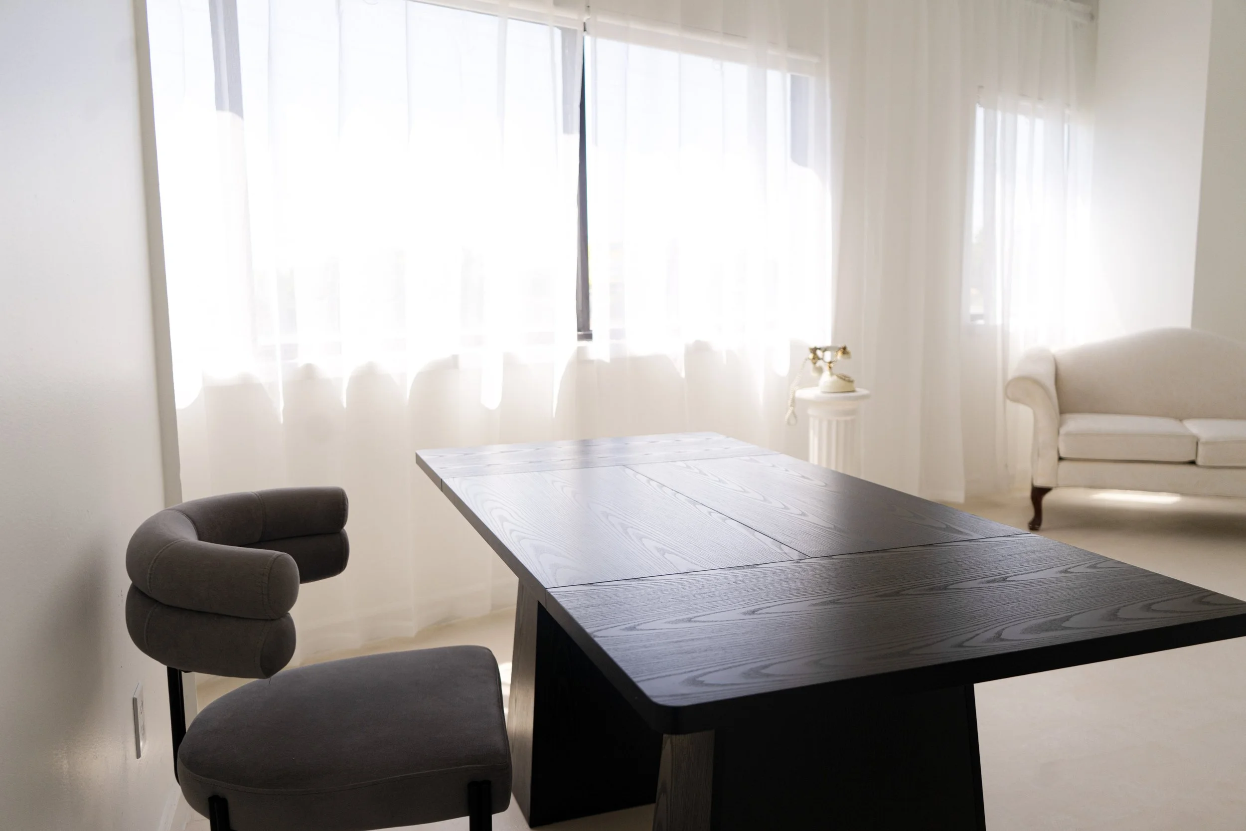 Minimalist dining room with a large dark wooden table, two taupe upholstered chairs, a white sofa, sheer white curtains, and a vintage rotary phone on a small table, all illuminated by natural light