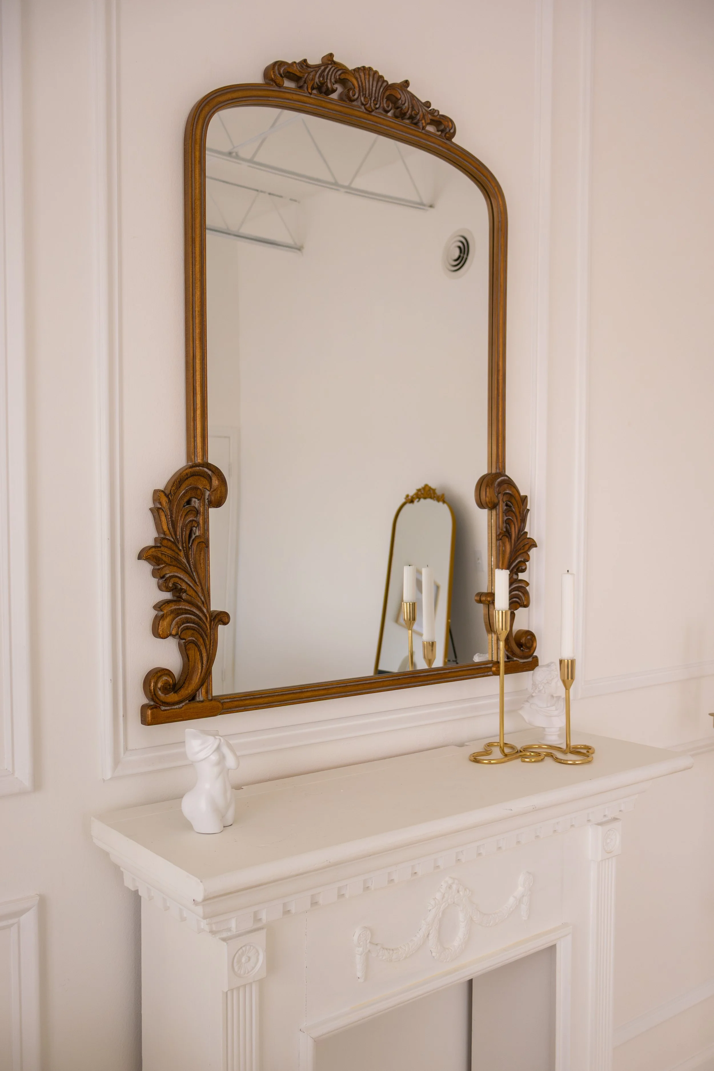 Decorative white fireplace mantel with a large ornate wooden mirror hanging above it. The mantel has a small white bunny sculpture and two gold candlestick holders with white candles. There is also a small white classical bust sculpture on the mantel. The room has white walls with architectural molding details.