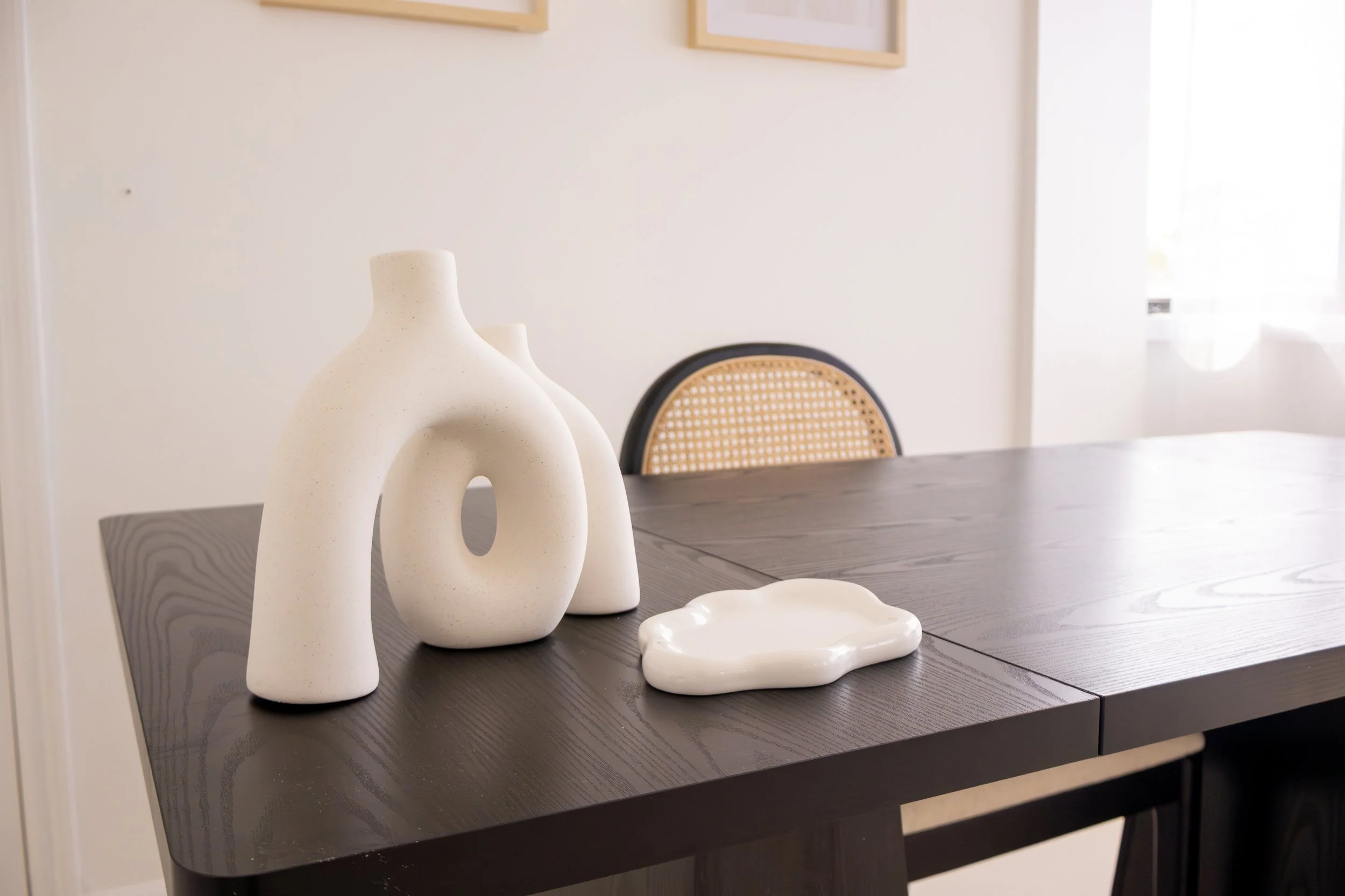 Decorative white ceramic vases and a white ceramic dish on a dark wooden dining table in a room with framed pictures and a window with white curtains