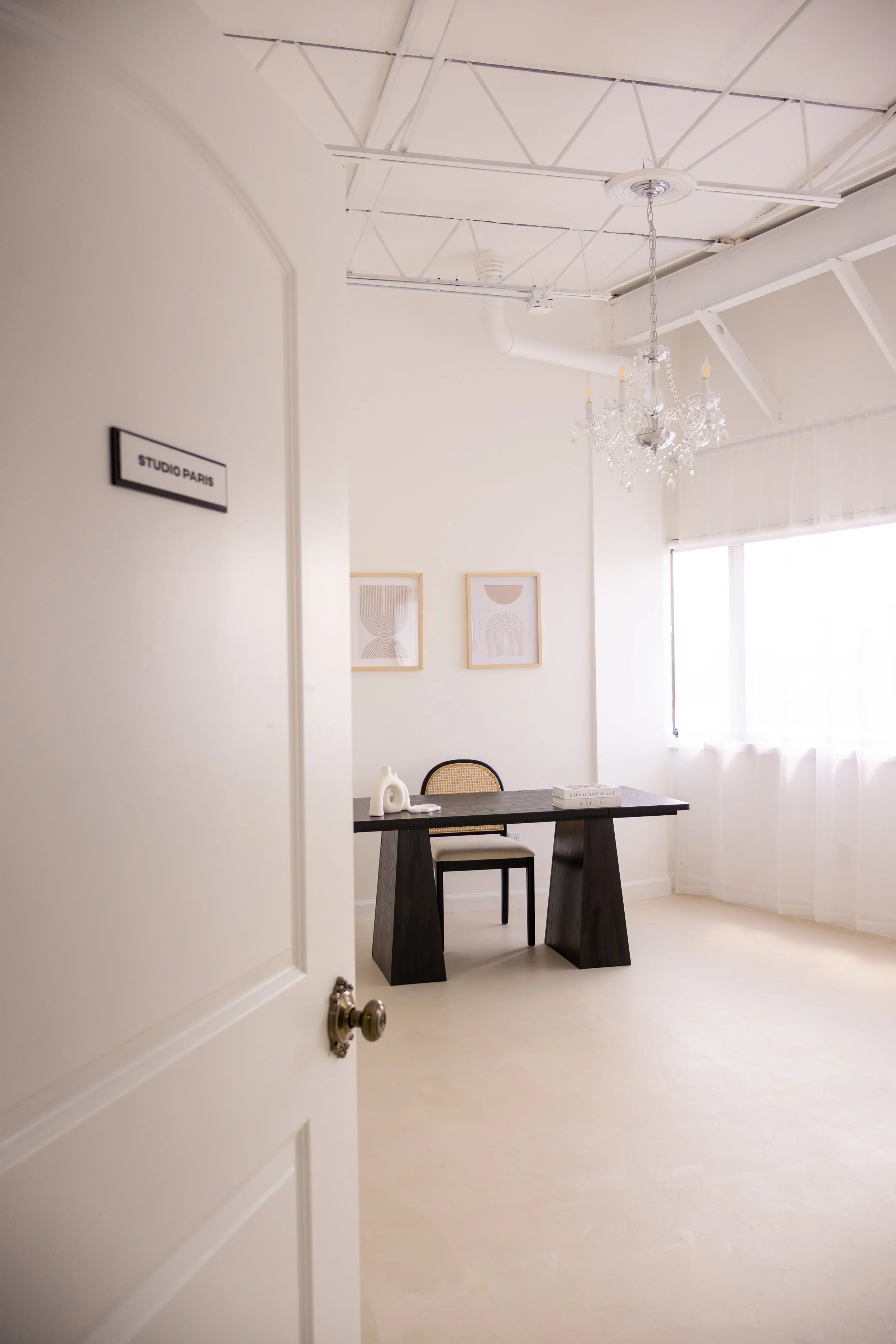 A minimalist office with a black desk, a chair, a white decorative sculpture, framed art on the wall, a chandelier, and a large window with sheer curtains, viewed through an open door labeled 'STUDIO PARIS'.