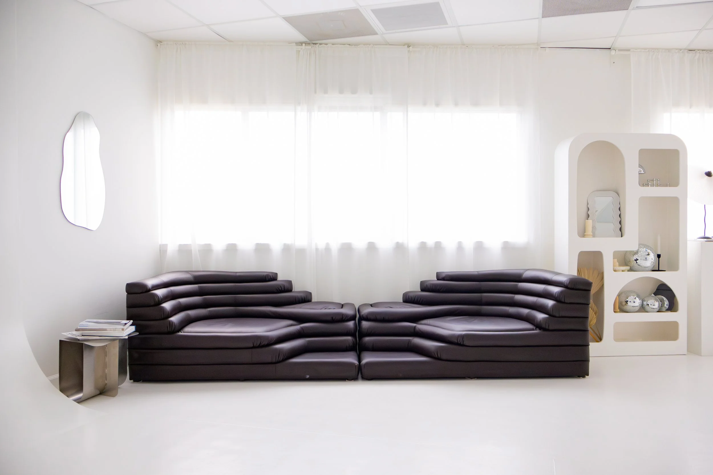 Minimalist living room with two black leather modern sofas facing each other, white walls, a small silver side table with books, and a white shelving unit decorated with vases, candles, and small decorations, large windows with sheer curtains, and a white wall mirror.