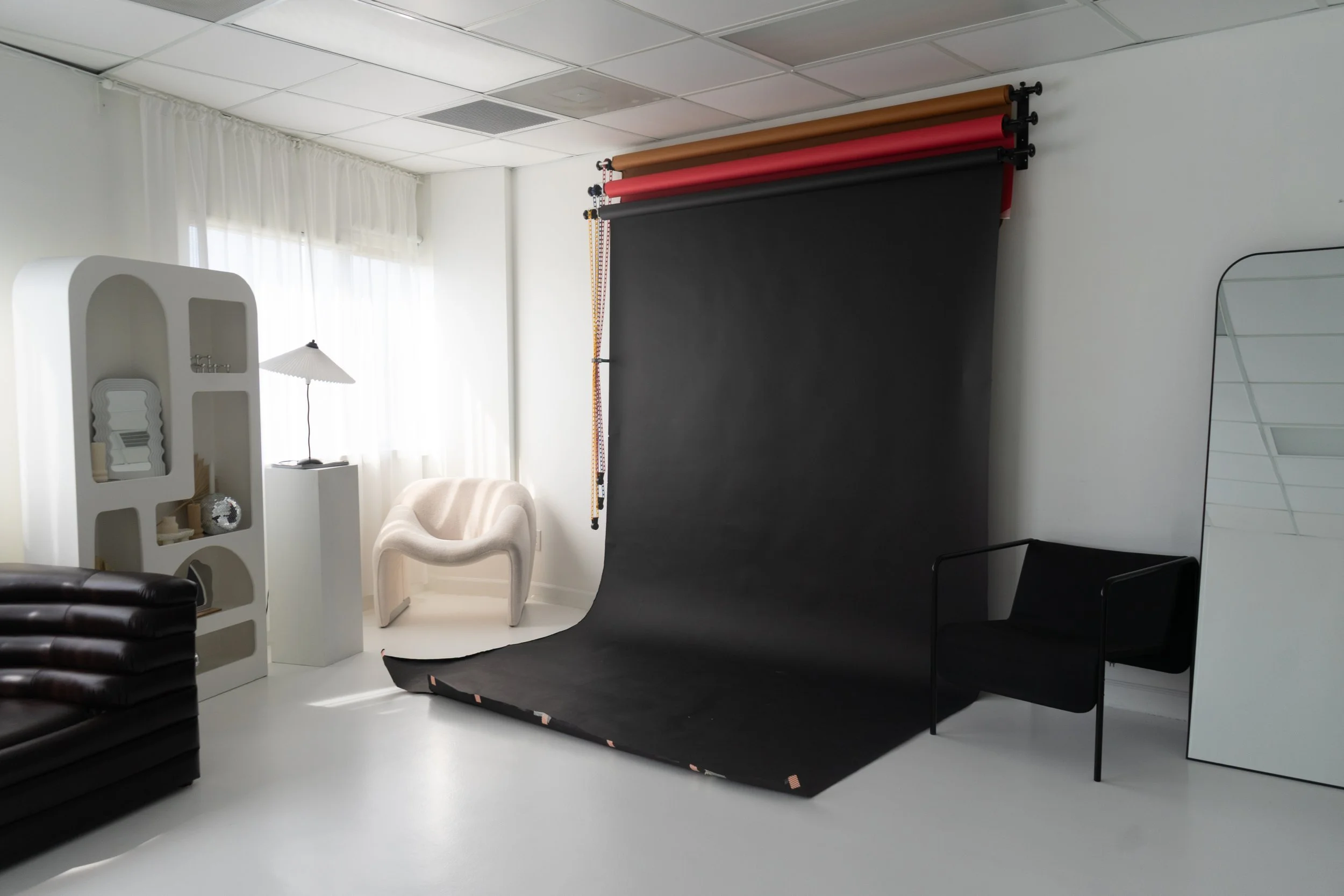 Photography studio with a black backdrop, chairs, a window with white curtains, and various studio equipment.