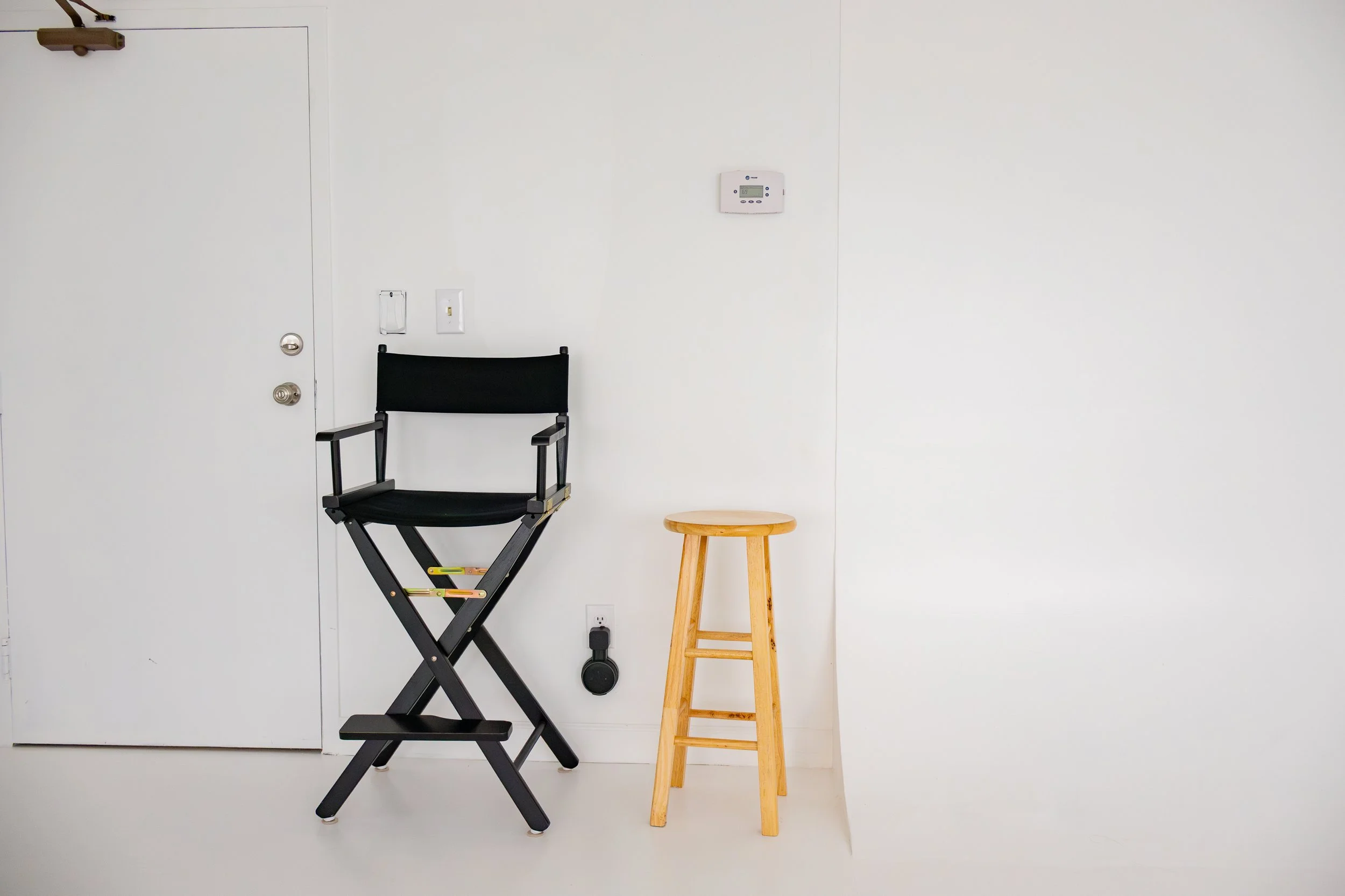 Black director's chair and a light wooden stool placed against a white wall near a door with a digital thermostat above and a power outlet with a plug below.