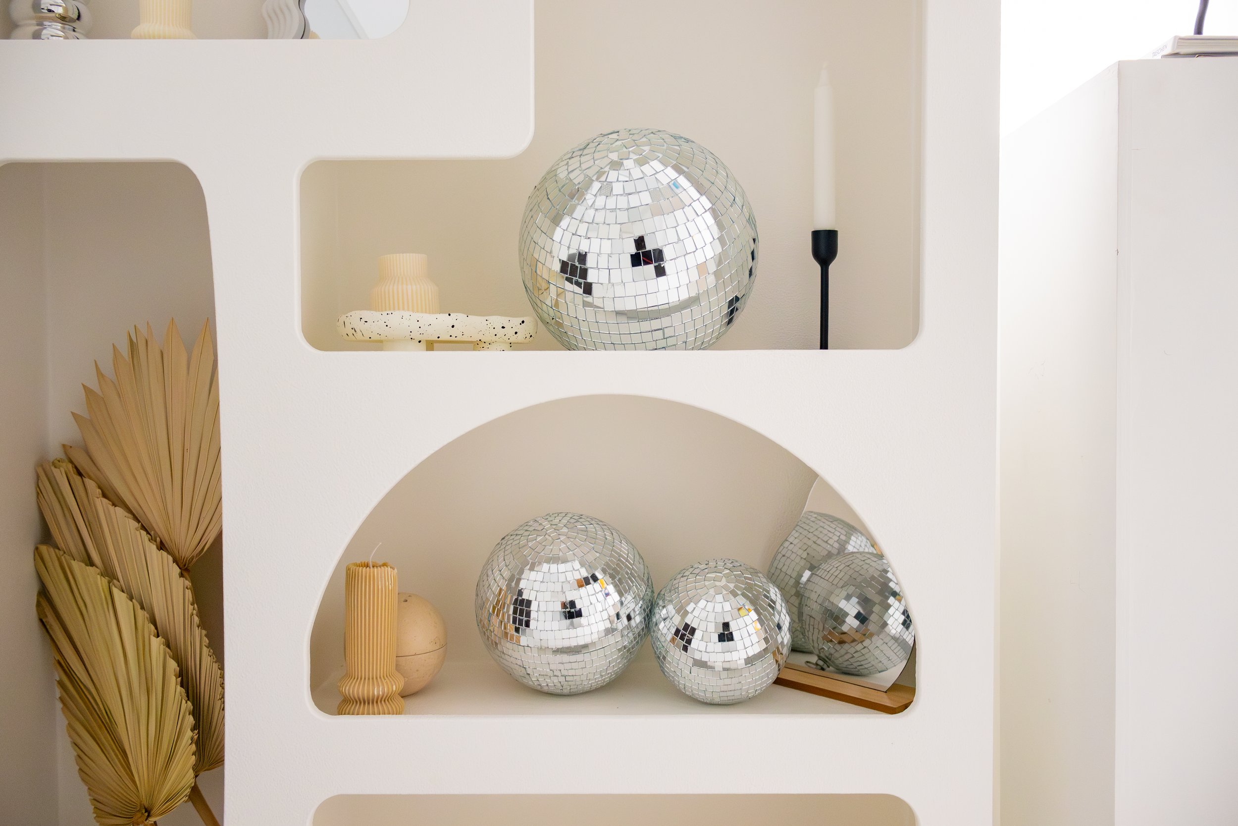 Decorative display shelf with small disco balls, dried palm leaves, candles, and minimalistic decor items in neutral tones.