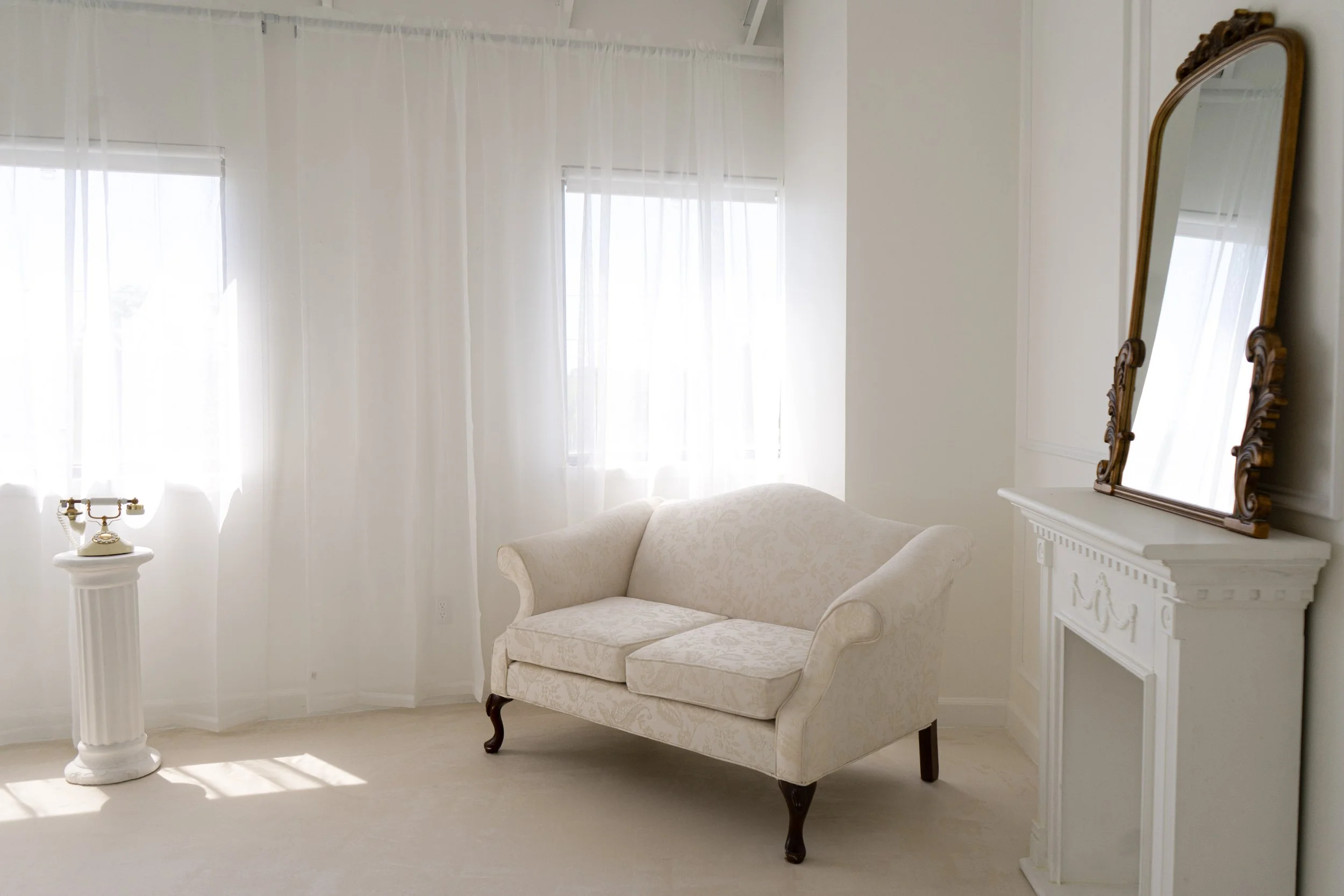 A white, vintage-style loveseat with wooden legs positioned in front of a window with sheer white curtains. To the right, a white fireplace mantel with an ornate wooden mirror reflection. To the left, a white pedestal with a vintage rotary phone. The room is bright with natural light.