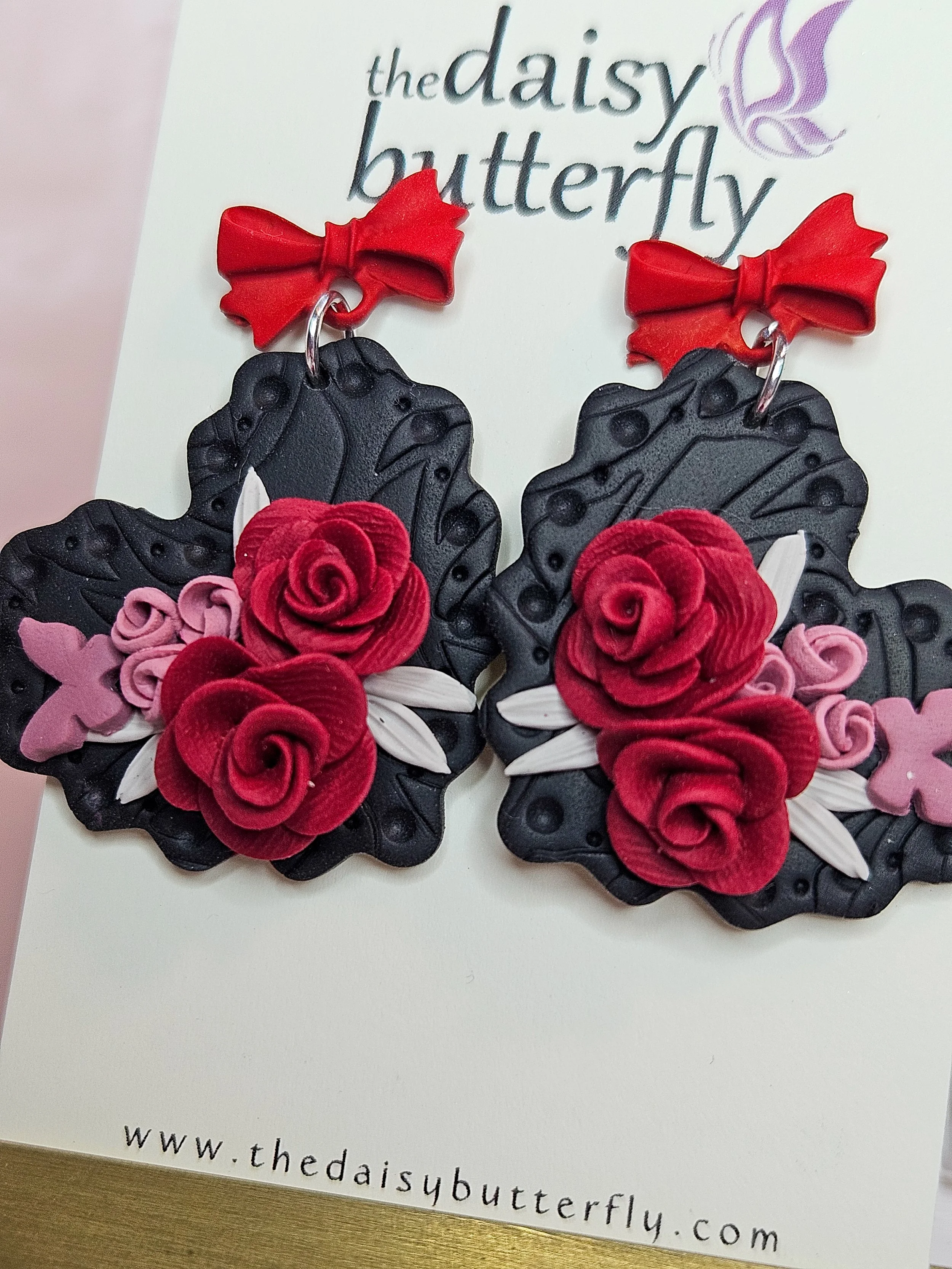 Pair of earrings with black butterfly-shaped bases decorated with red roses, pink butterflies, and white leaves, topped with red bows made of ribbon.