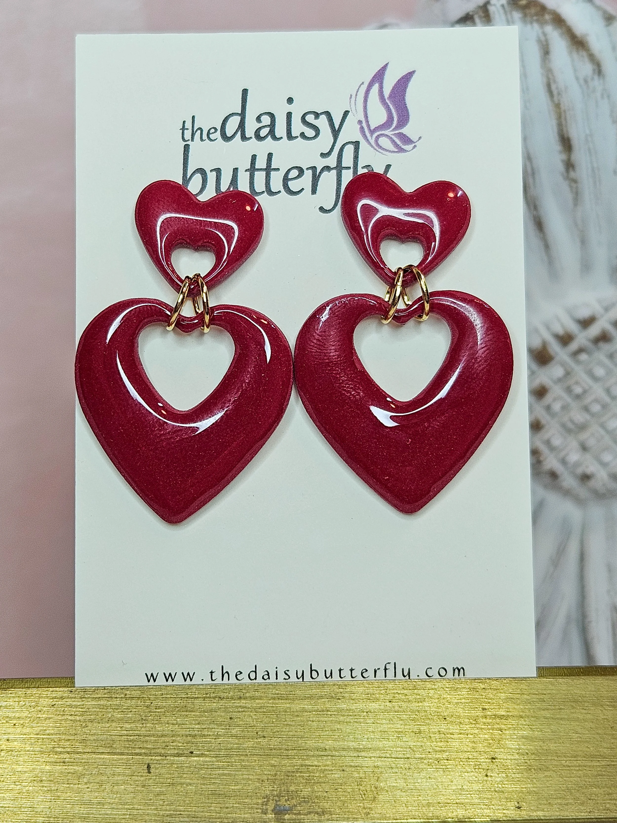 Red heart-shaped earrings with smaller open hearts inside, displayed on a card with the logo 'the daisy butterfly' and website address.