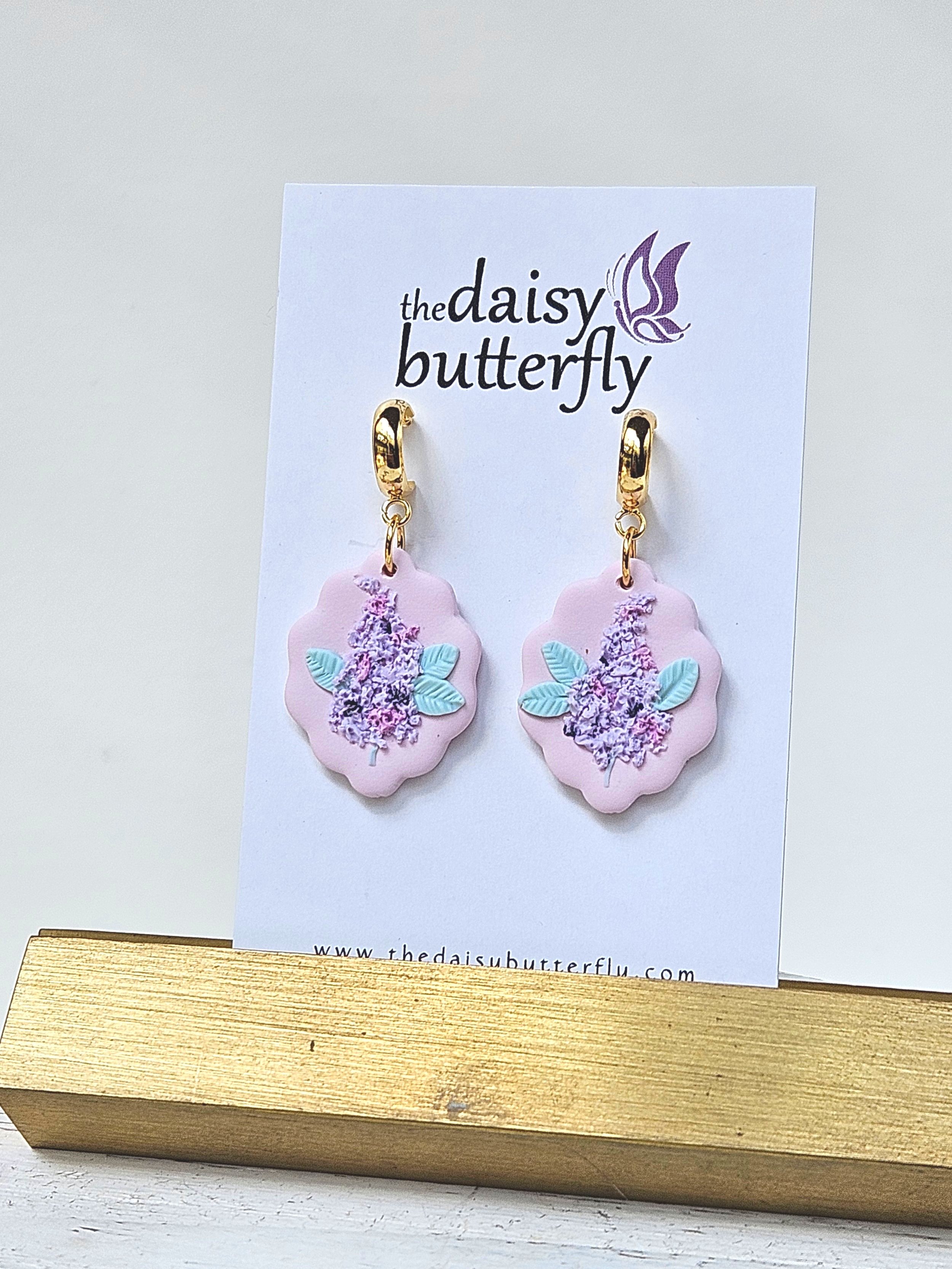 A pair of handmade pastel lilac earrings with textured purple flower clusters and mint green leaves on a scalloped lavender base, attached to gold huggie hoops.