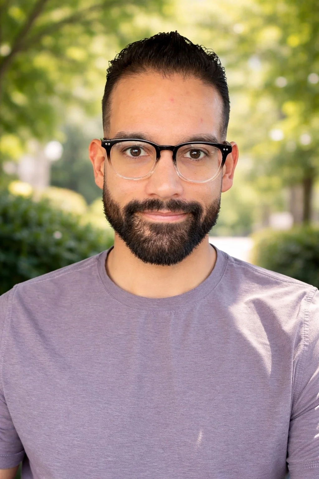 A man (Daniel Gama) with dark hair, glasses, and a beard standing outdoors with trees and greenery in the background.