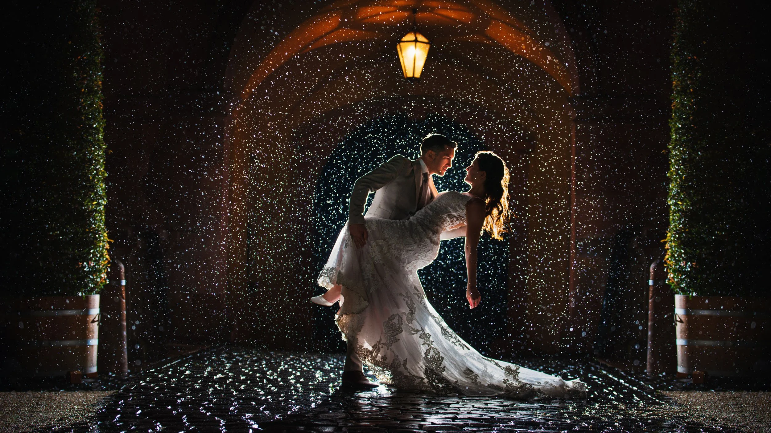 Bride and groom dancing under glowing lights in a cobblestone alleyway with gentle rain falling, symbolizing romantic wedding photography moments.
