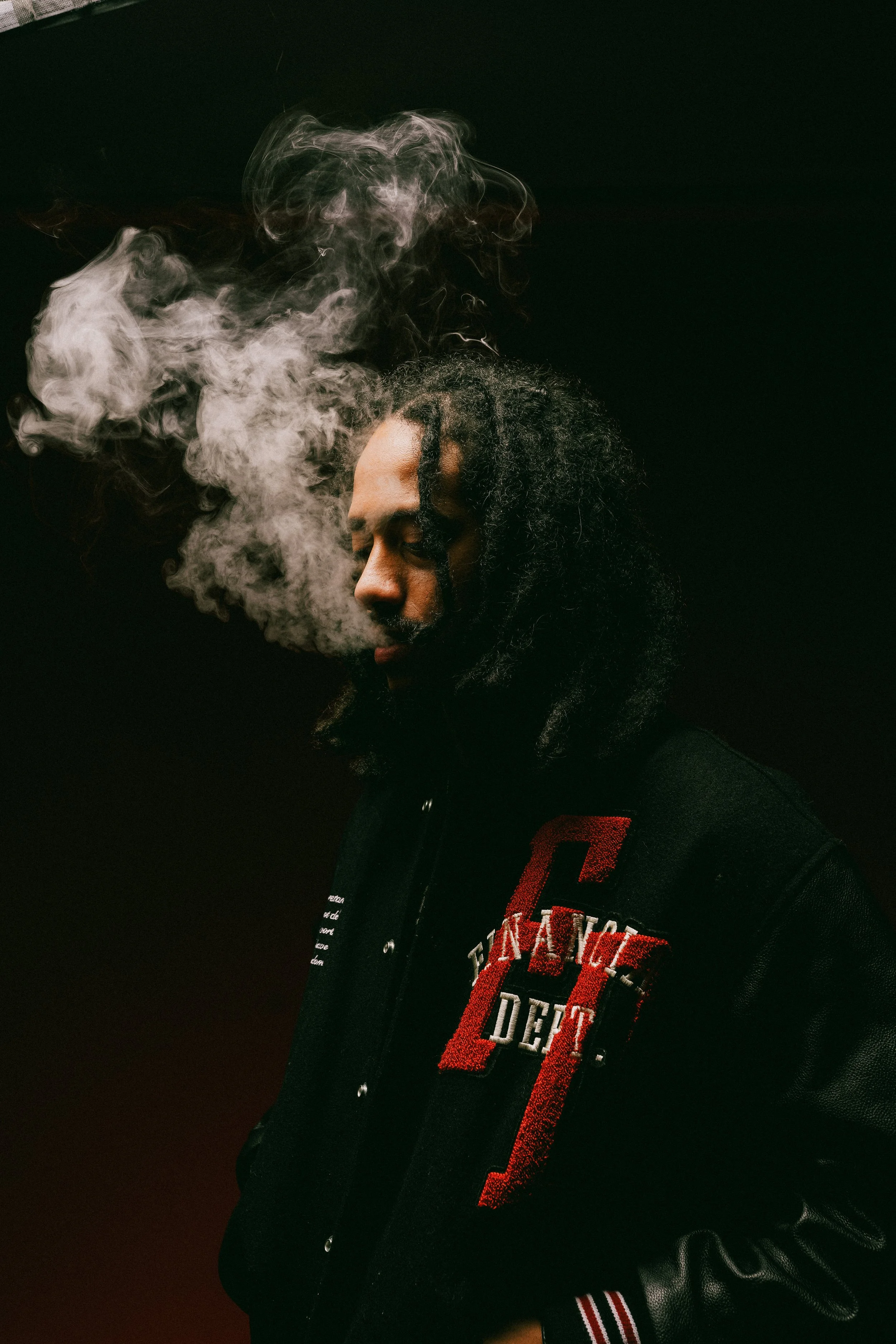 Person exhaling smoke in dim lighting, wearing a black jacket with red lettering.