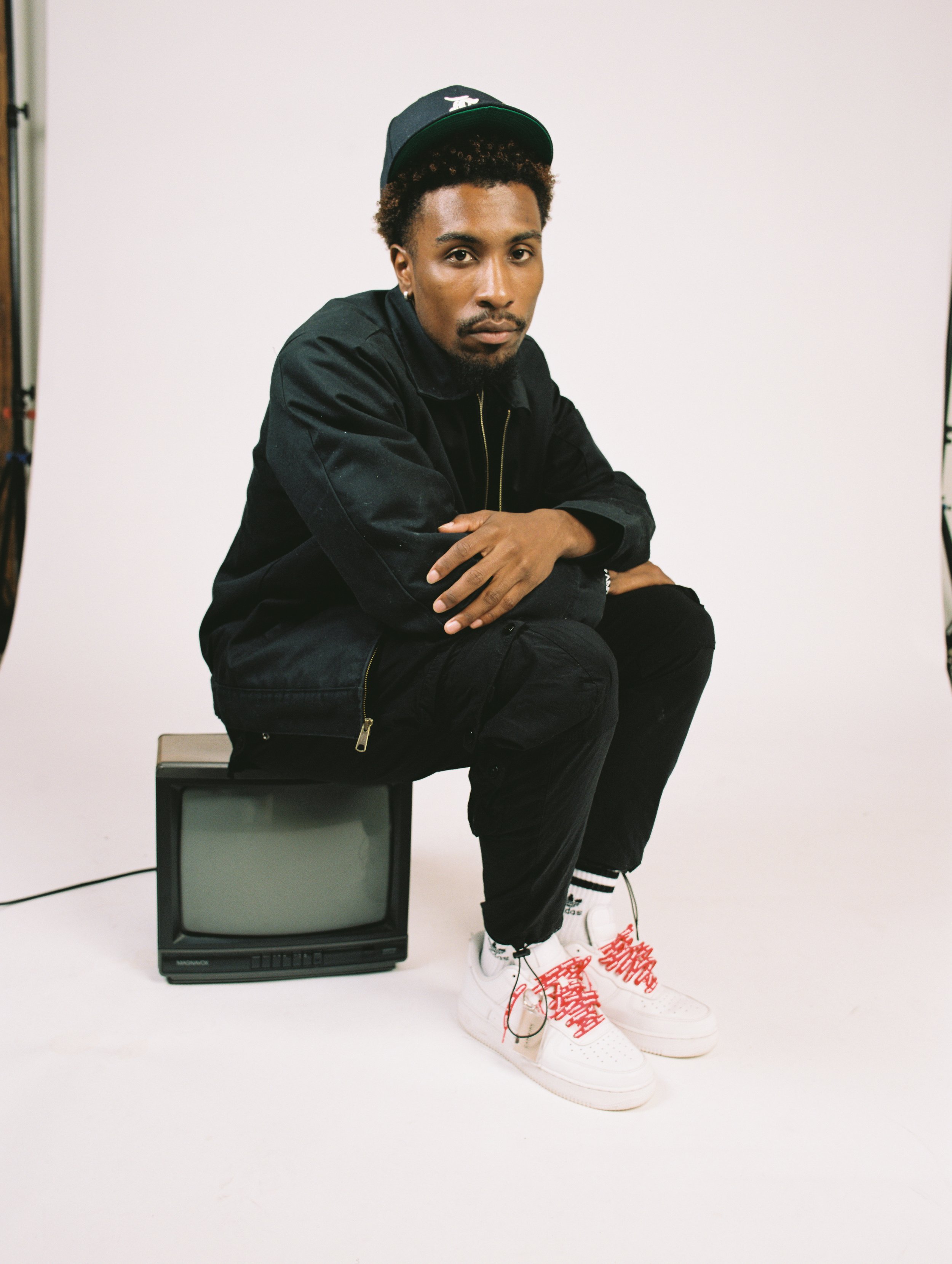 Person wearing black jacket and cap sitting on a vintage TV, white sneakers with red laces.