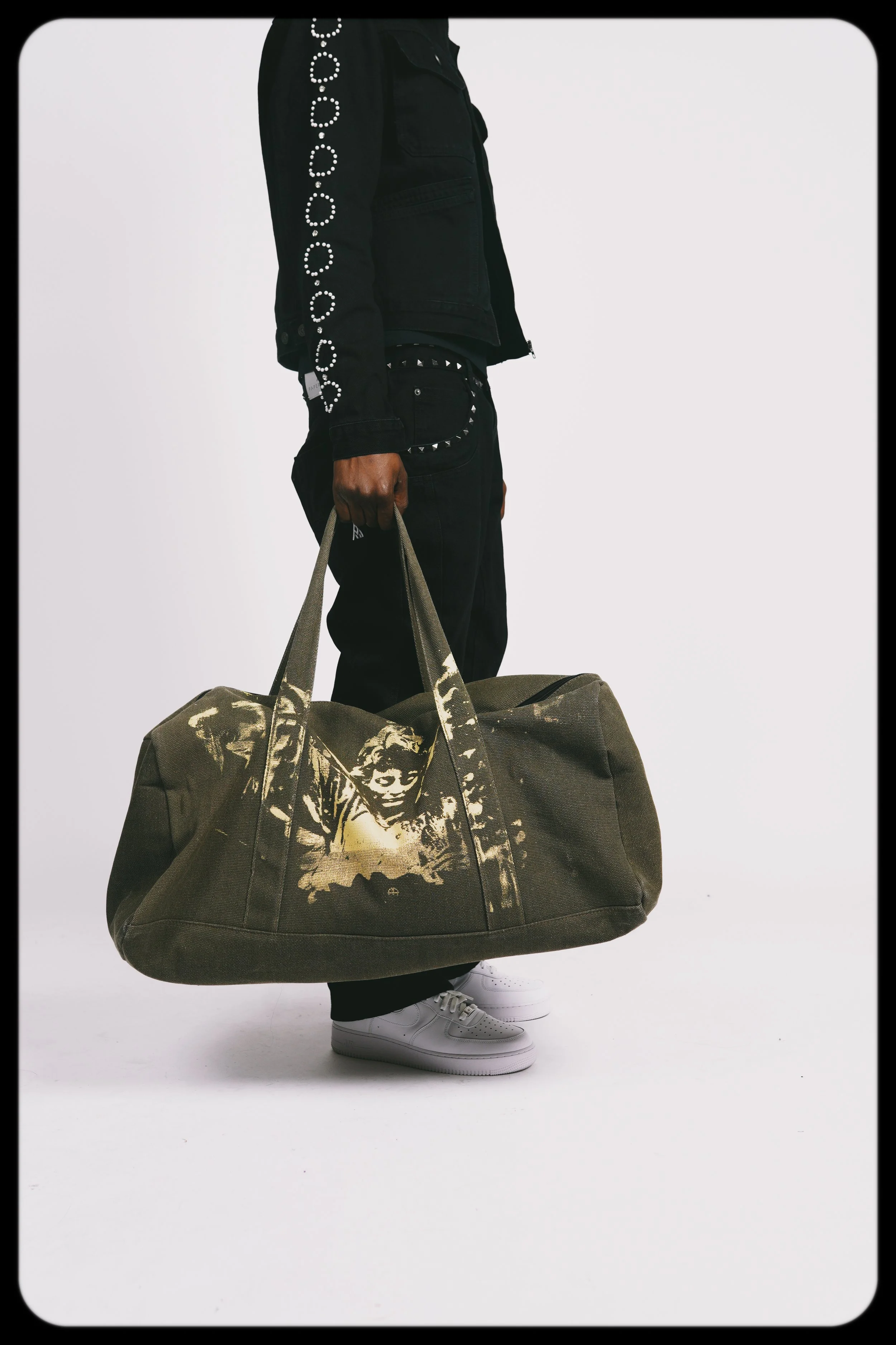 Person holding a large olive green duffel bag with a yellow graphic design, wearing dark pants with studs and white sneakers.