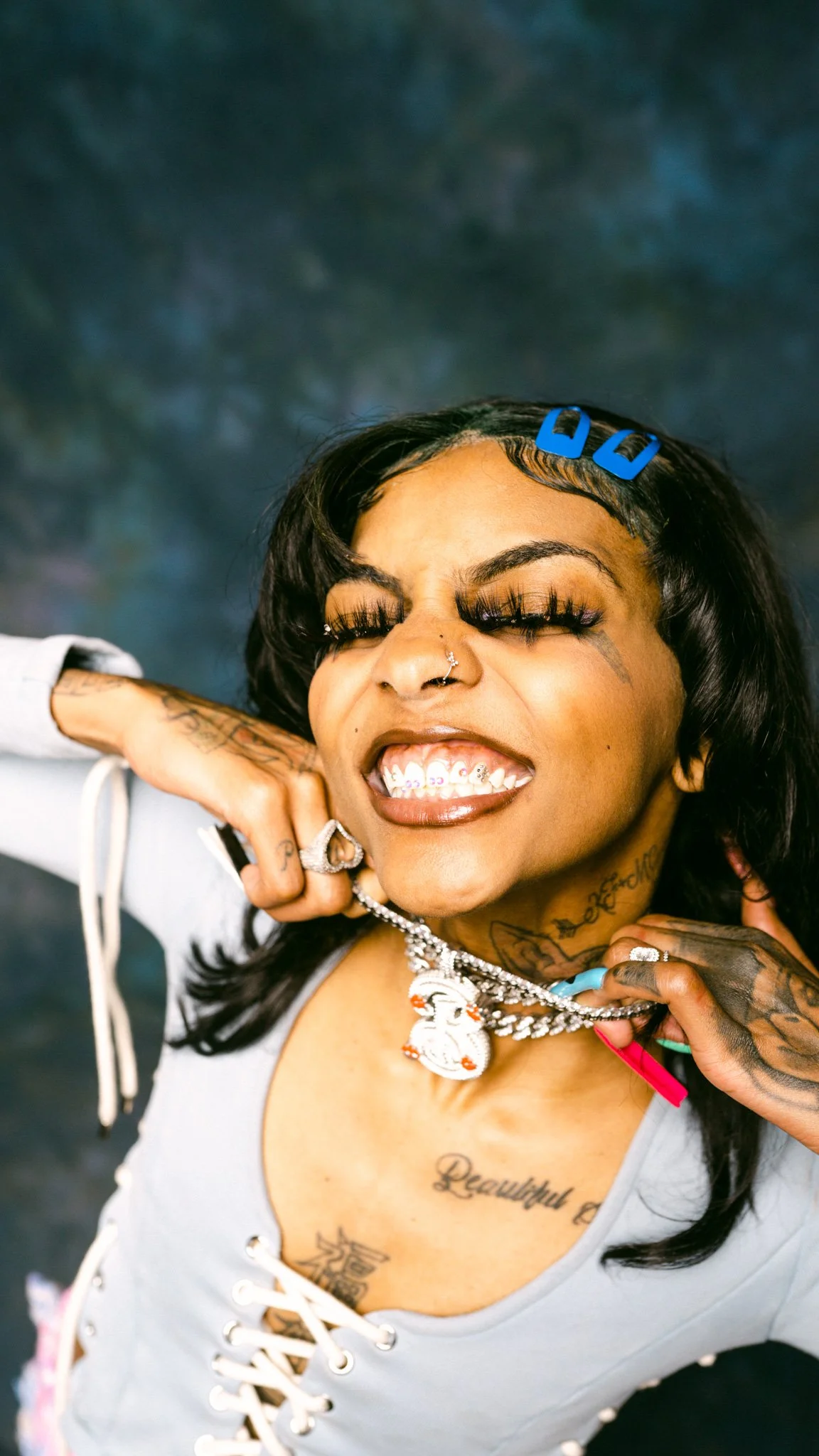 Smiling woman with tattoos, wearing jewelry and blue hair clips, showing teeth with designs.