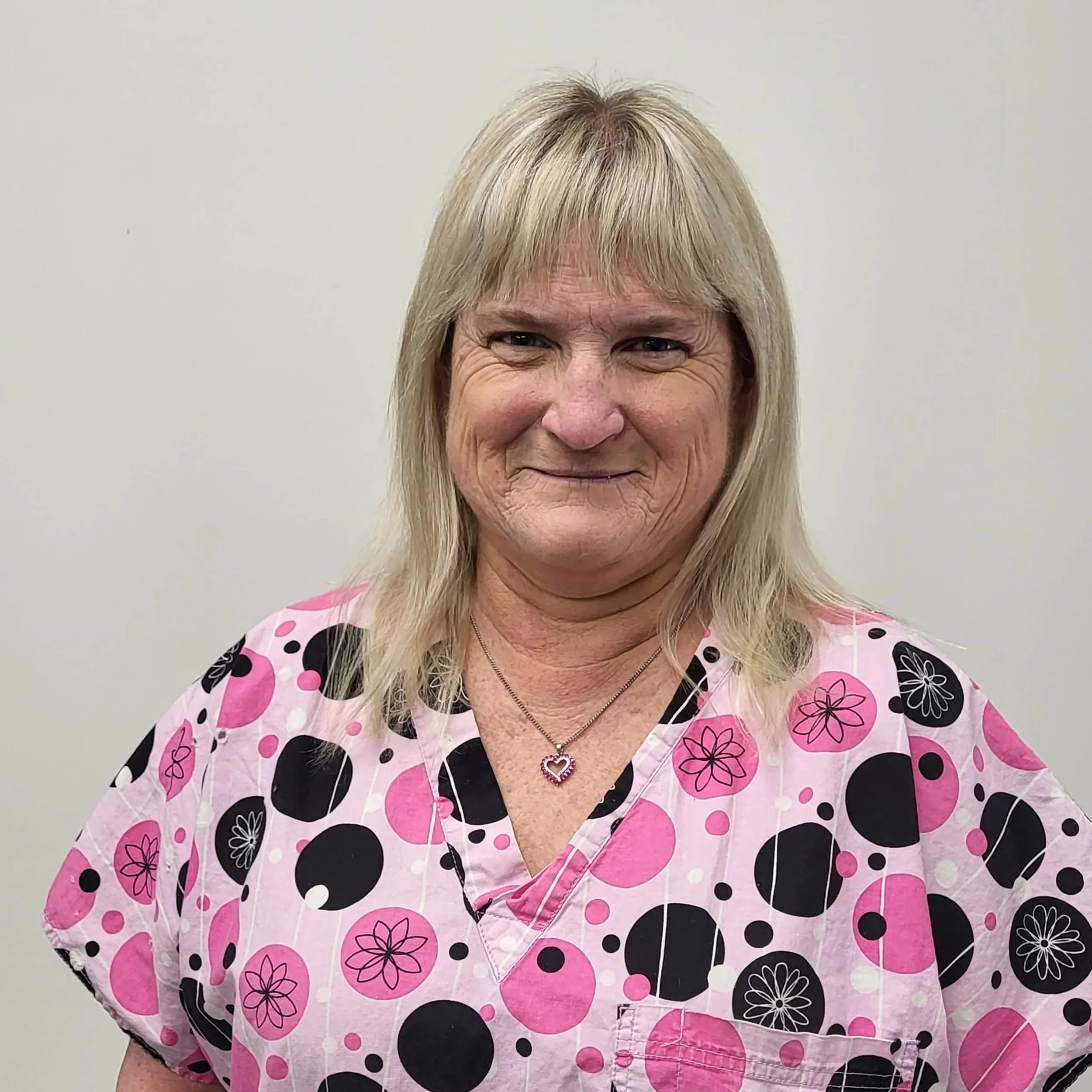 Jo Kantor wearing a pink and black patterned scrub top, standing against a plain background. The scrub top features circular and floral designs.