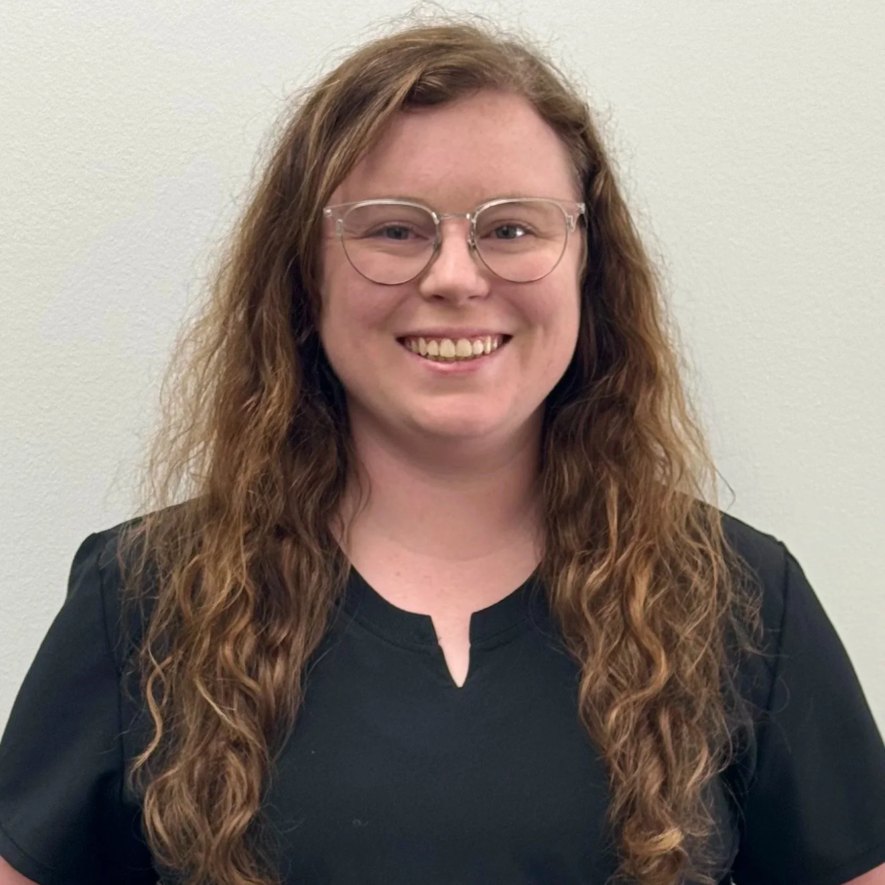 Samantha , with long, wavy hair and glasses, is smiling and wearing a black shirt against a plain background.