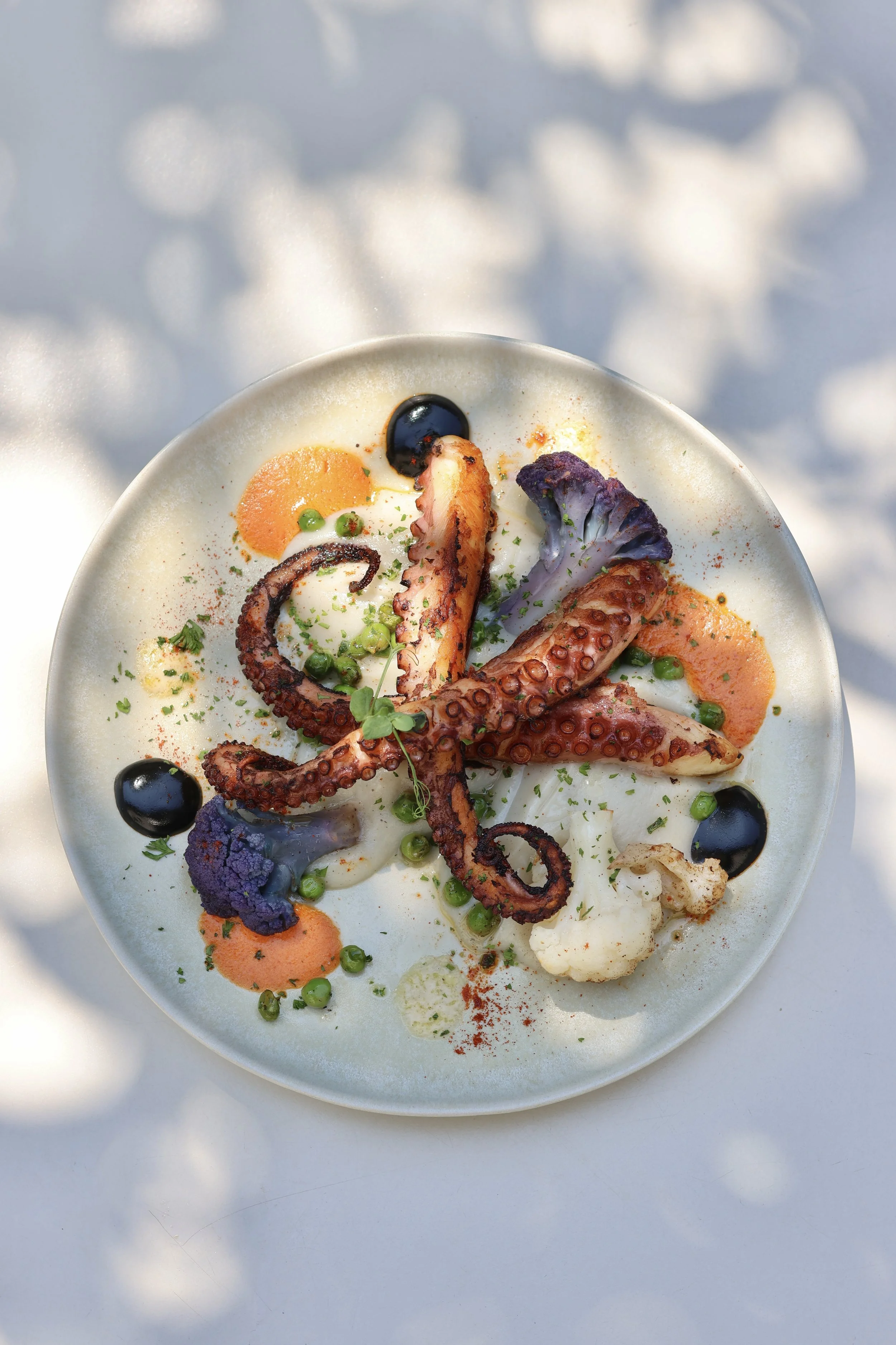 Plat gastronomique avec pieuvre grillée, légumes violets, cauliflower, olives noires, sauce orange, herbes, et épices sur une assiette blanche.