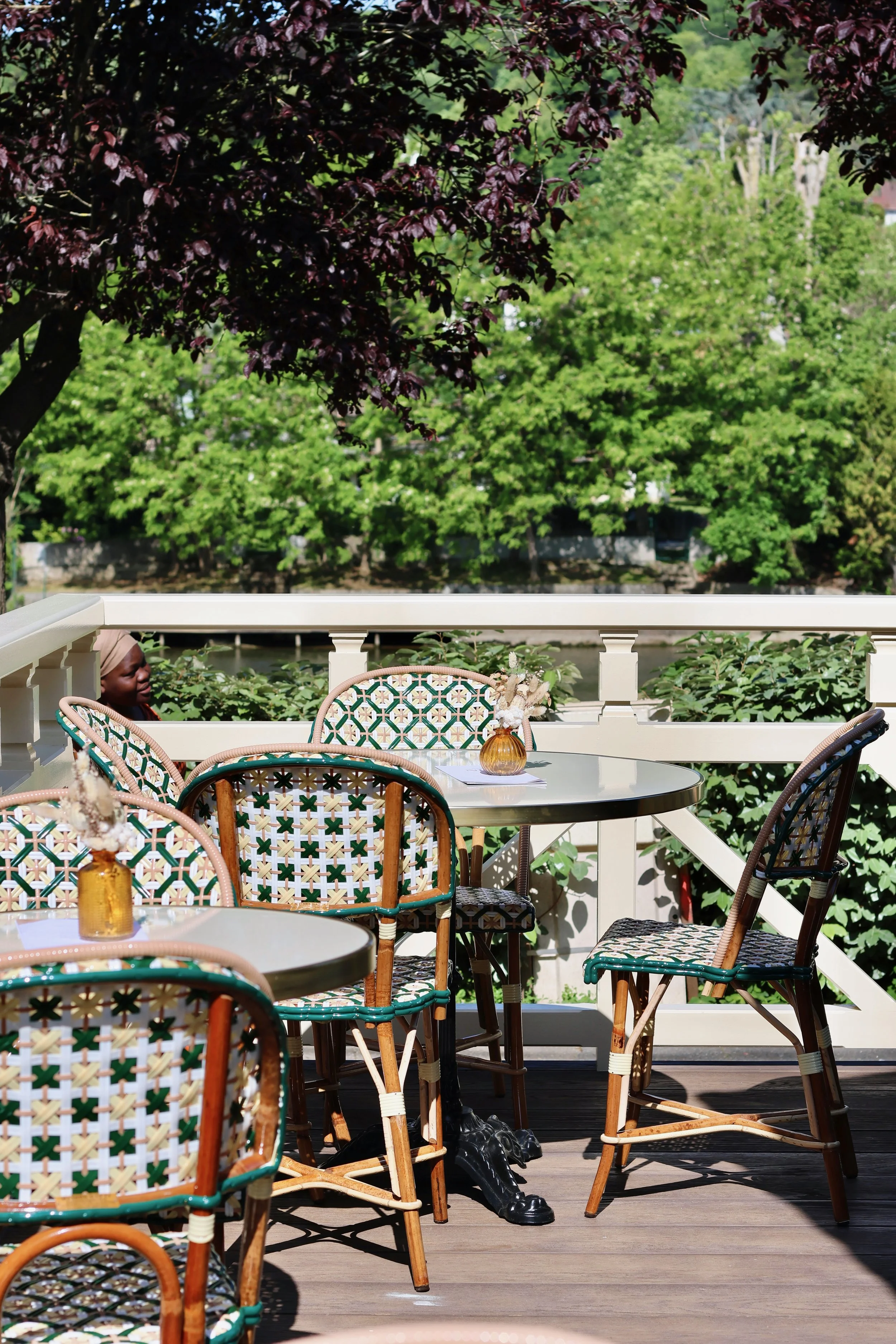 Une terrasse extérieure avec des chaises en rotin et un tableau rond, avec des vases en verre contenant des fleurs, entourée de verdure et d'arbres en plein air.