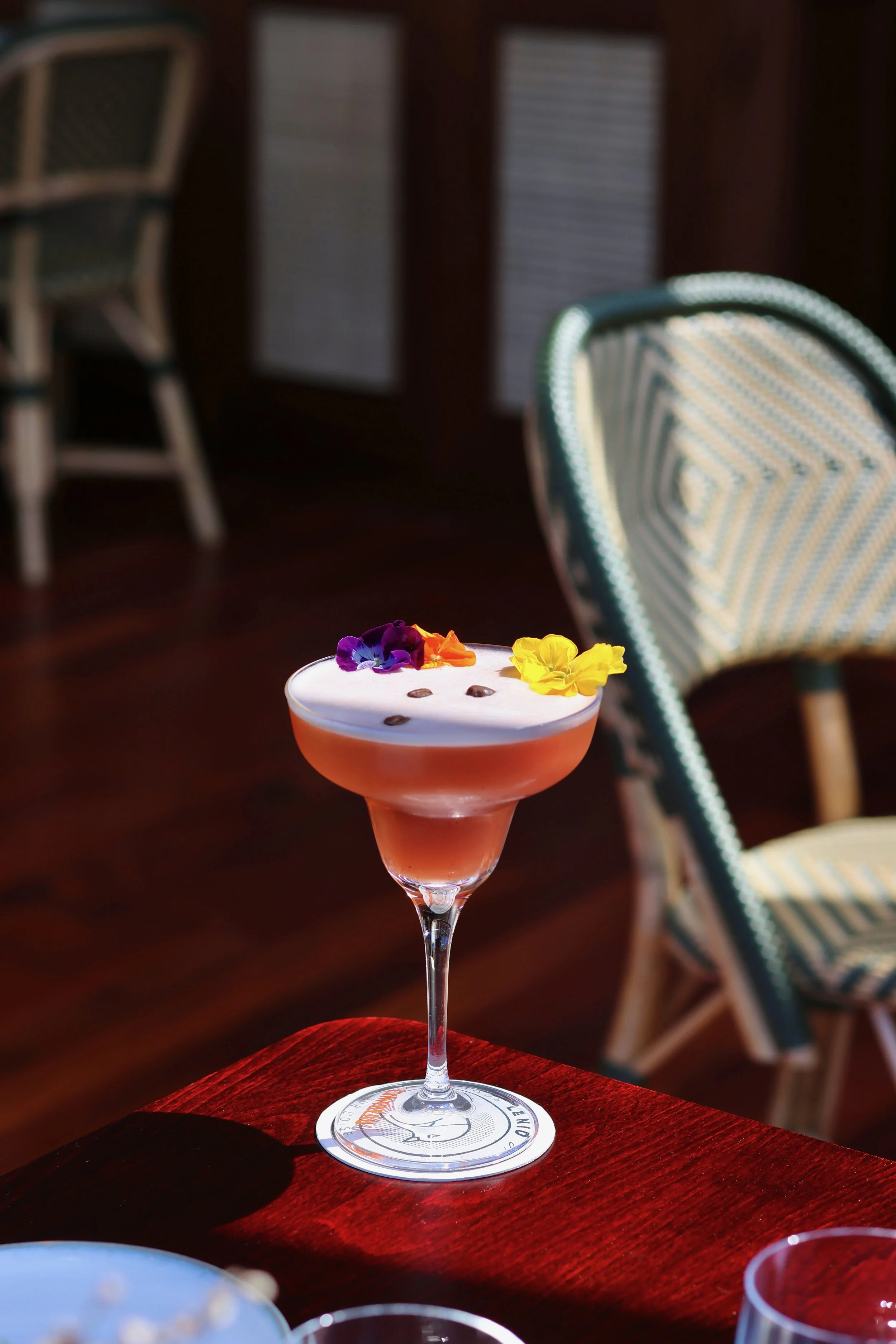 Un cocktail rose dans un verre à pied, décoré avec des fleurs violettes, jaunes et orange, et quelques grains de café en surface, posé sur une table en bois.