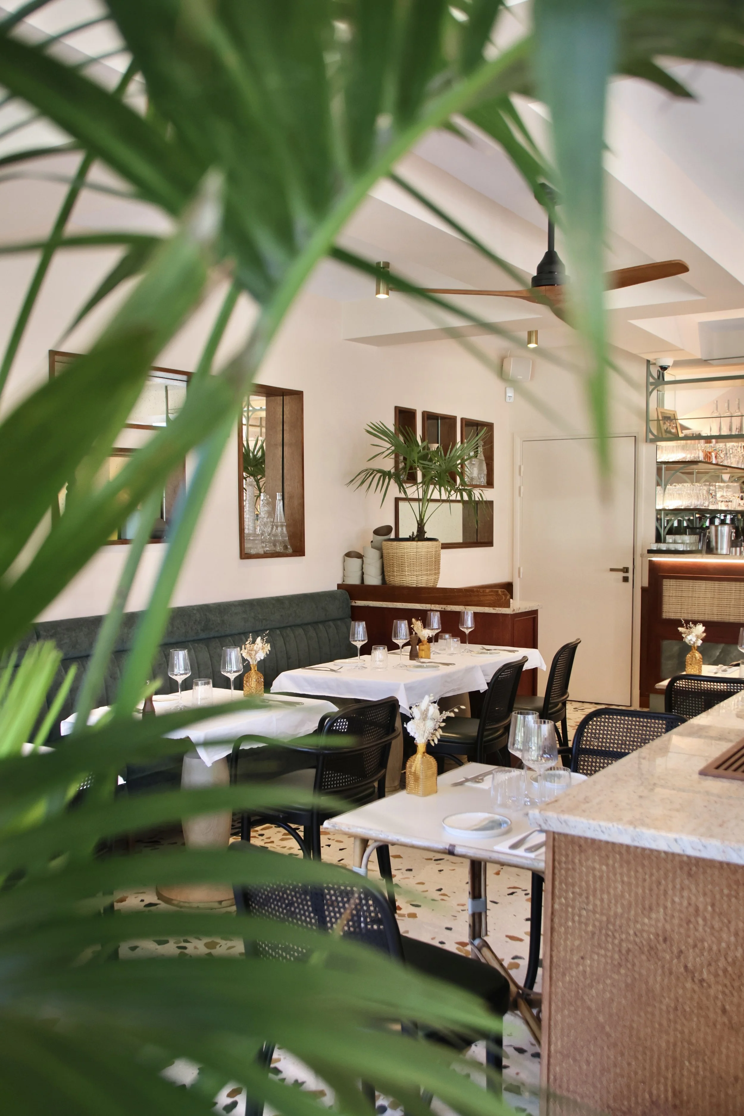 Intérieur d'un restaurant élégant avec des tables dressées avec des verres à vin et des bouquets de fleurs, vue partielle à travers des feuilles de plante verte.