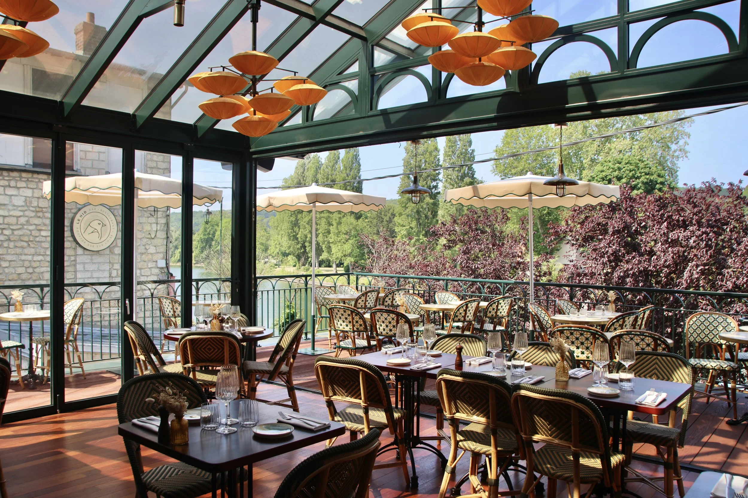 Salle de restaurant avec tables et chaises en intérieur et terrasse extérieure ombragée avec parasols blancs, vue sur la nature avec arbres verts, décor sophistiqué et lumière naturelle.