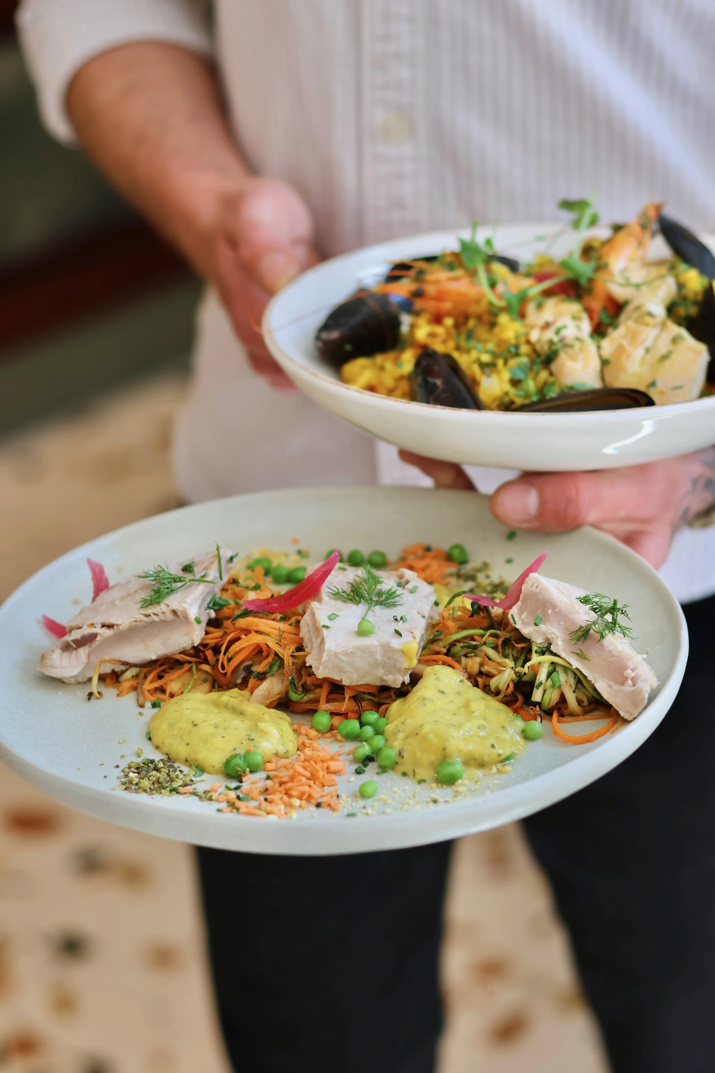 Deux plats de nourriture présentés, une personne tient un plat contenant un riz coloré avec des fruits de mer et des herbes, et une autre personne tient un plat avec des morceaux de poulet, des légumes râpés, des pois verts, une sauce jaune, et diver