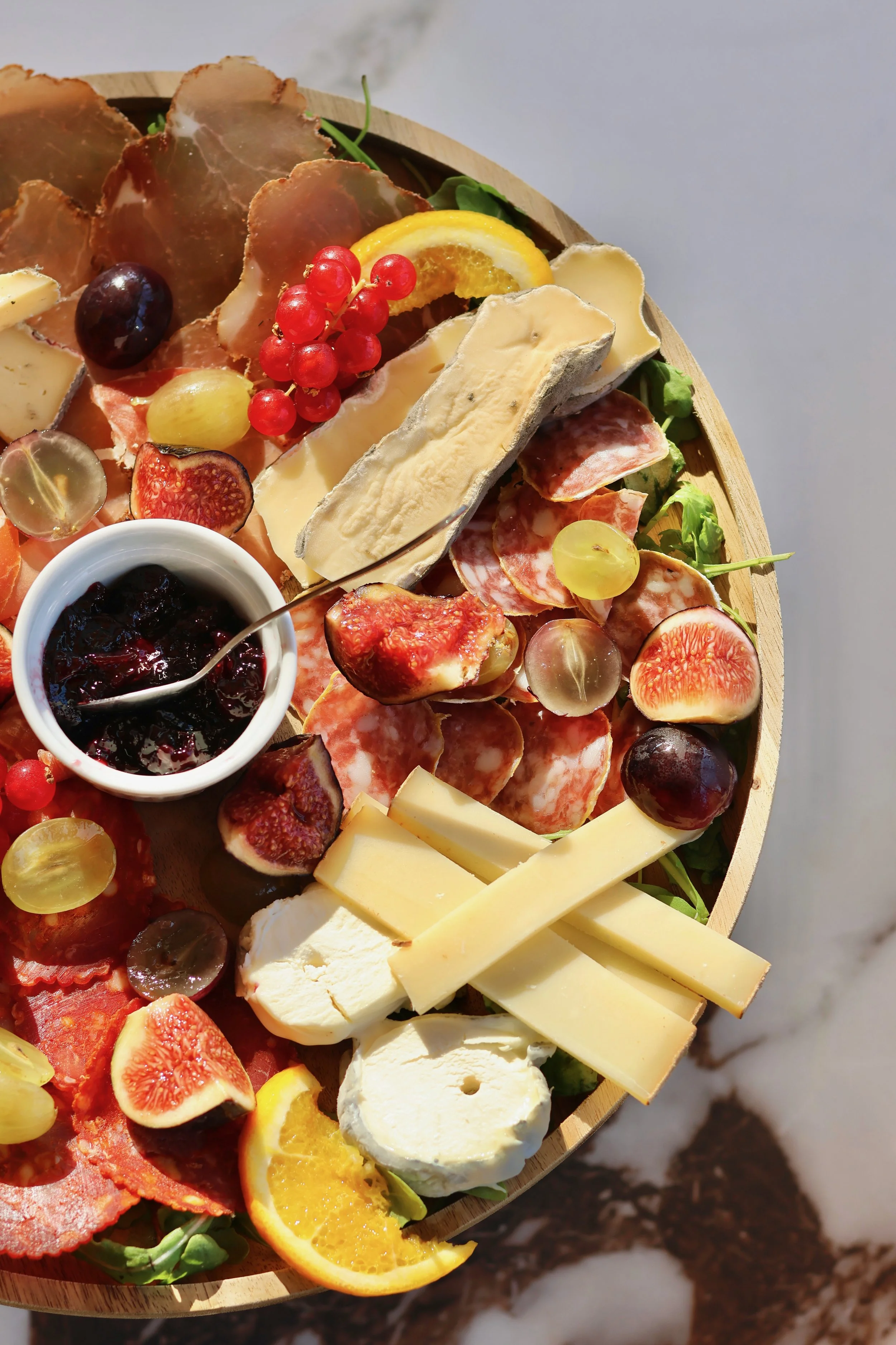 Plate de fromages, fruits, charcuterie, et confiture, avec tranches de fromage, figues, raisins, oranges, et autres accompagnements sur un plateau en bois.
