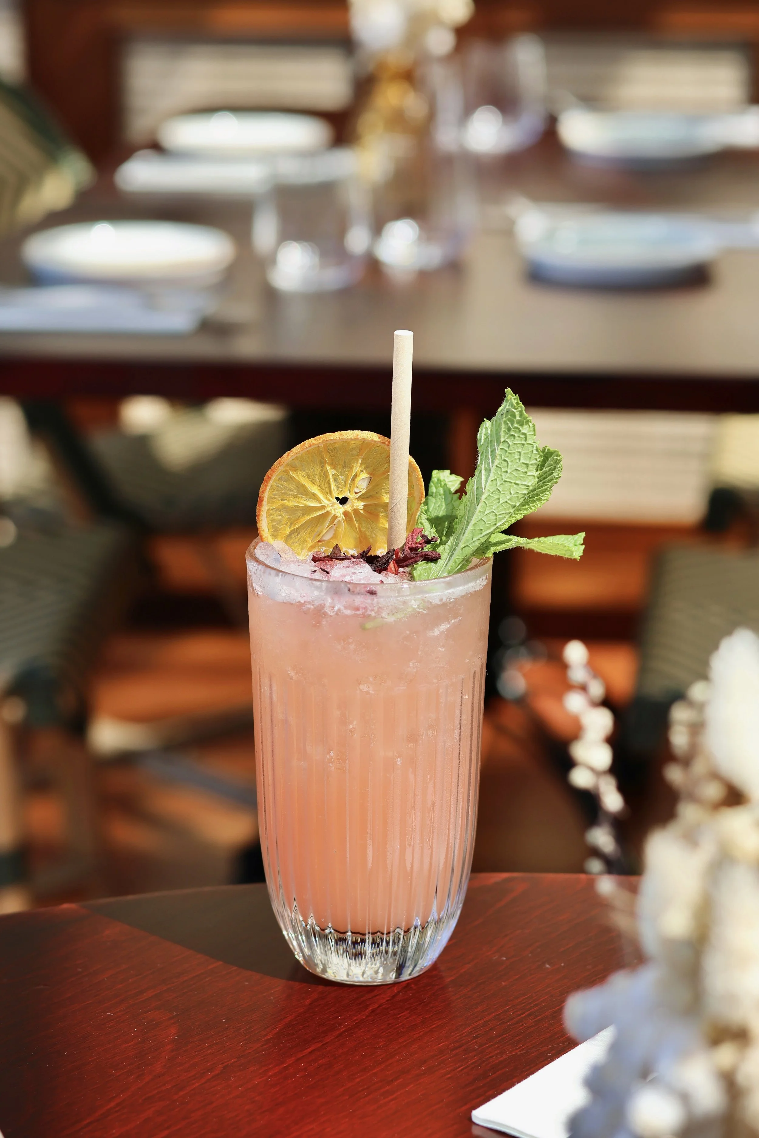 Un verre long avec une boisson rose garnie de tranche de citron, feuilles de menthe et un bâtonnet en bois, placé sur une table en bois.