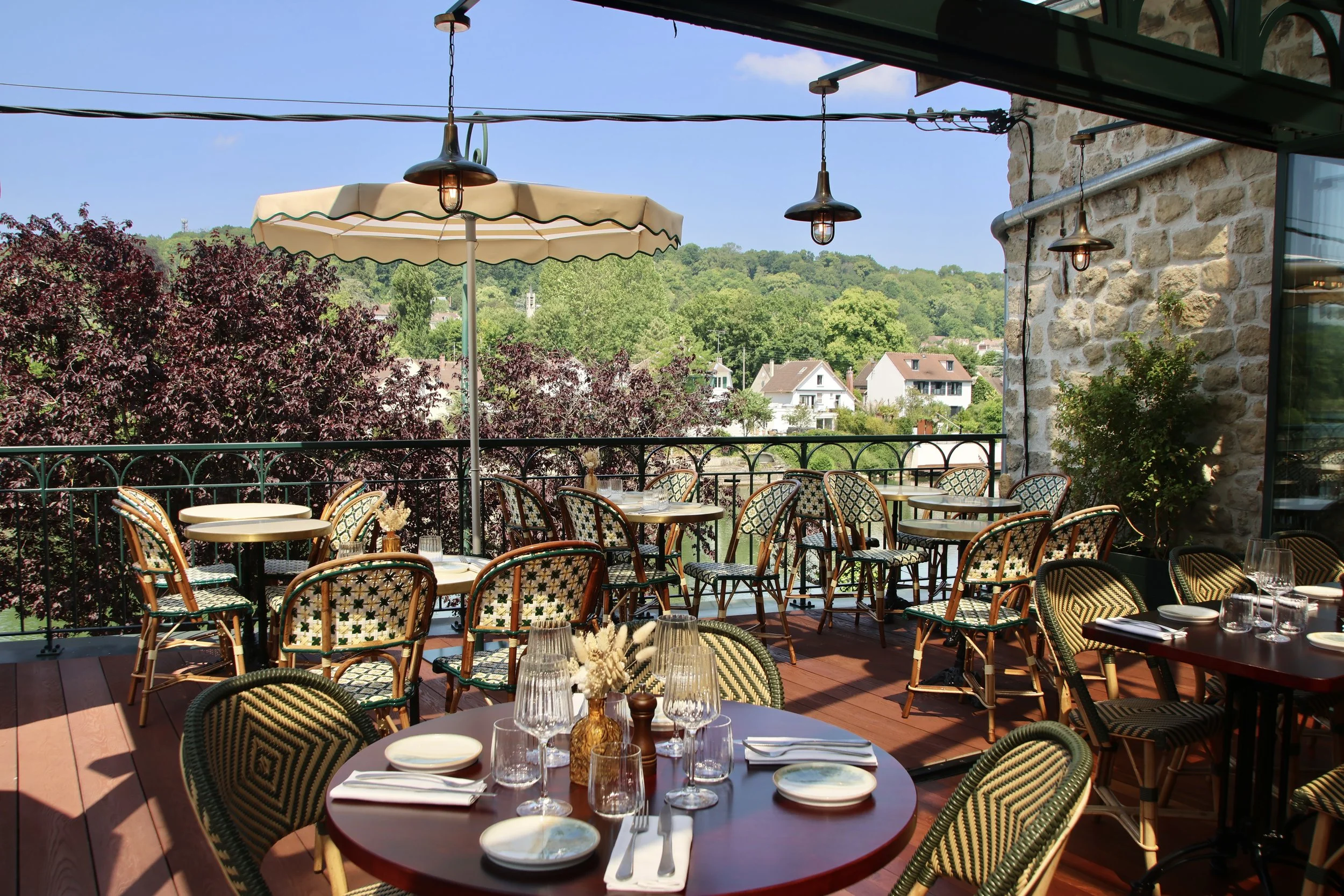 Terrasse de restaurant en plein air avec tables, chaises, parasol, et vue sur un paysage verdoyant avec des maisons et des collines.