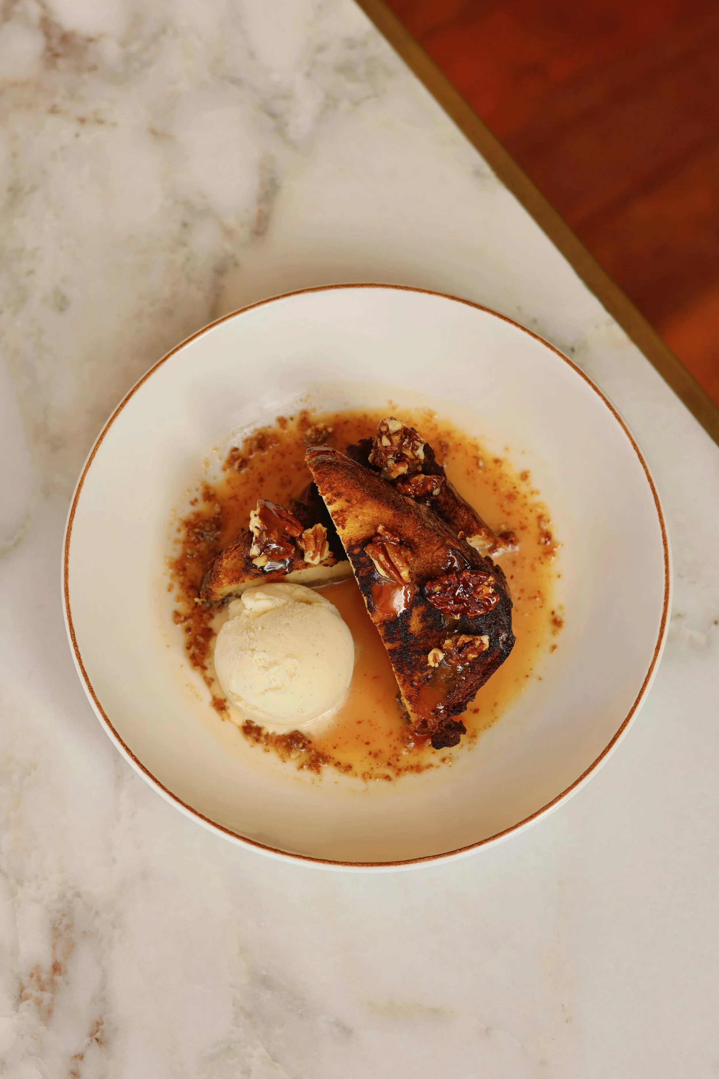 Dessert avec une tranche de gâteau au caramel recouverte de noix, une boule de glace à côté, dans un bol blanc sur une table en marbre Blanco.