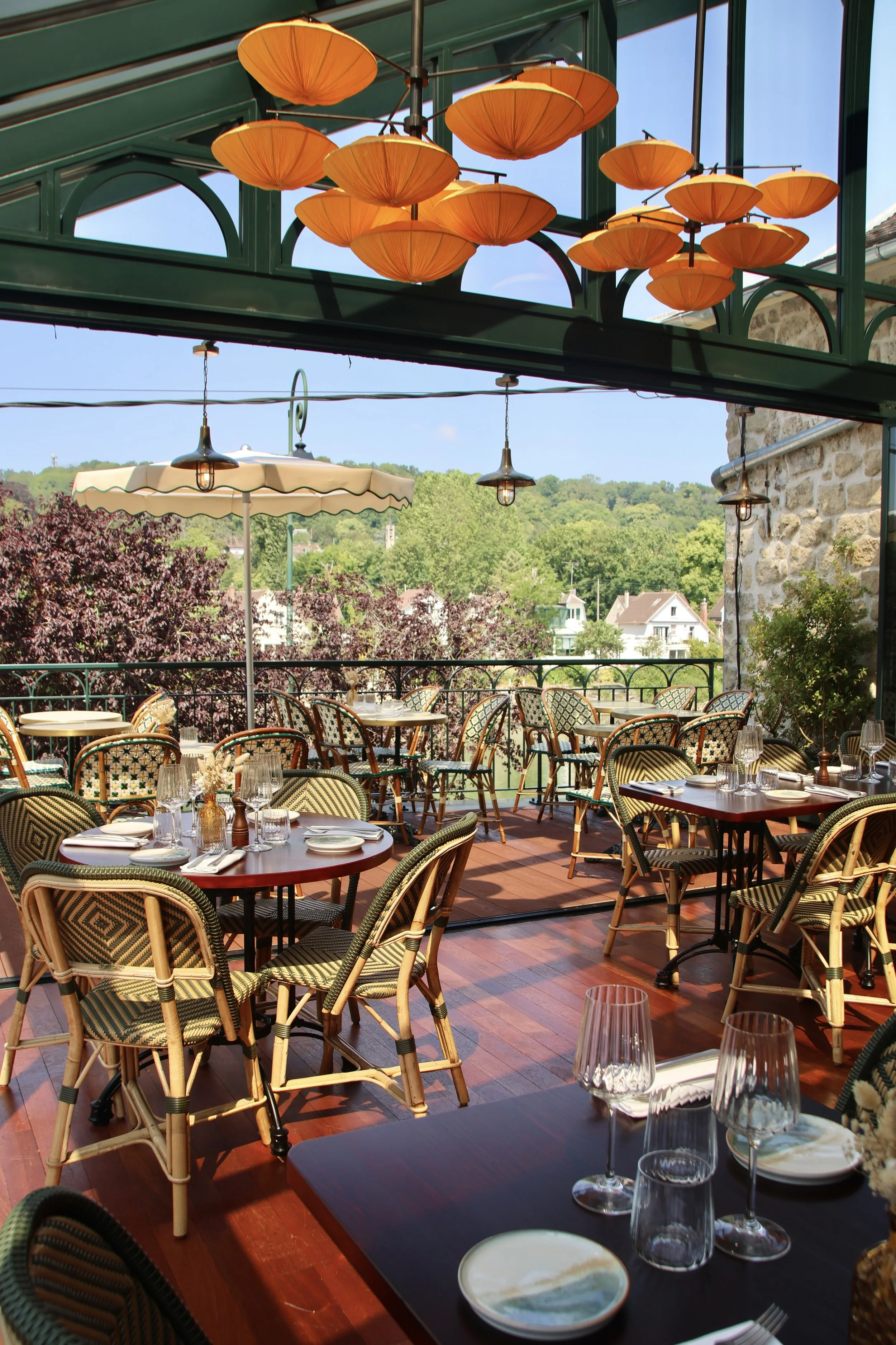 Terrasse de restaurant en plein air avec tables dressées, chaises en rotin, parasol, et un ciel bleu avec des arbres et des maisons en arrière-plan.