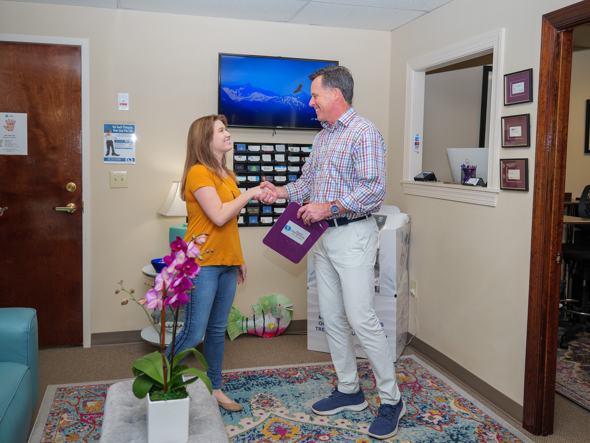 A woman in a yellow shirt is shaking hands with a man in a plaid shirt holding a folder in an office setting. There are informational posters on the wall, a TV displaying a mountain landscape, and a purple orchid on a table in the foreground.