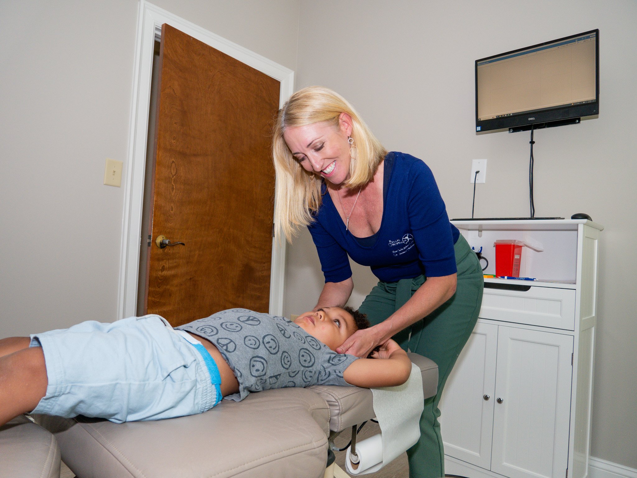 A child lying on a chiropractic treatment table while a female chiropractor gently holds the child's neck in a clinic setting. The room has a wooden door, a wall-mounted computer screen, and a cabinet with medical supplies.