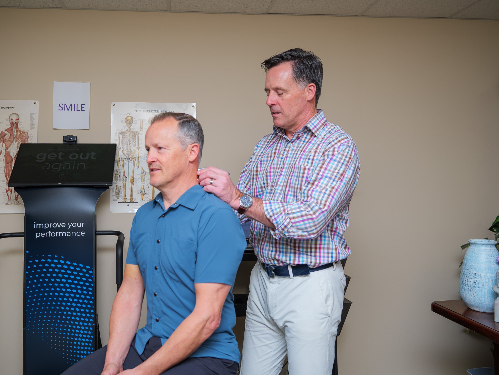 A physical therapist assessing a seated man's shoulder in a clinic setting.