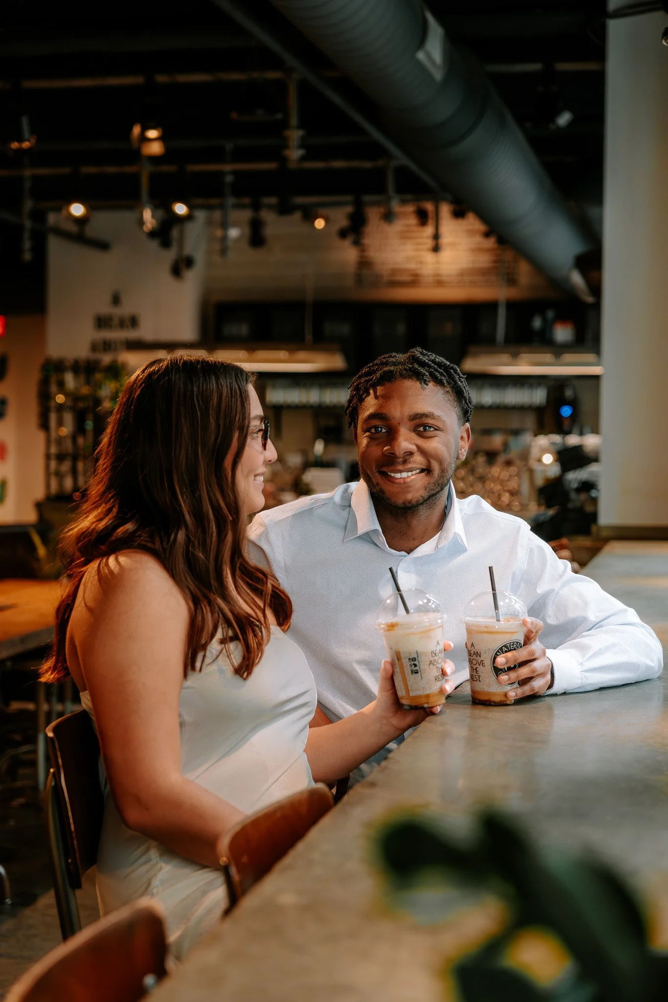 A couple sitting at a coffee shop, holding iced drinks, with a warm and inviting atmosphere in the background.