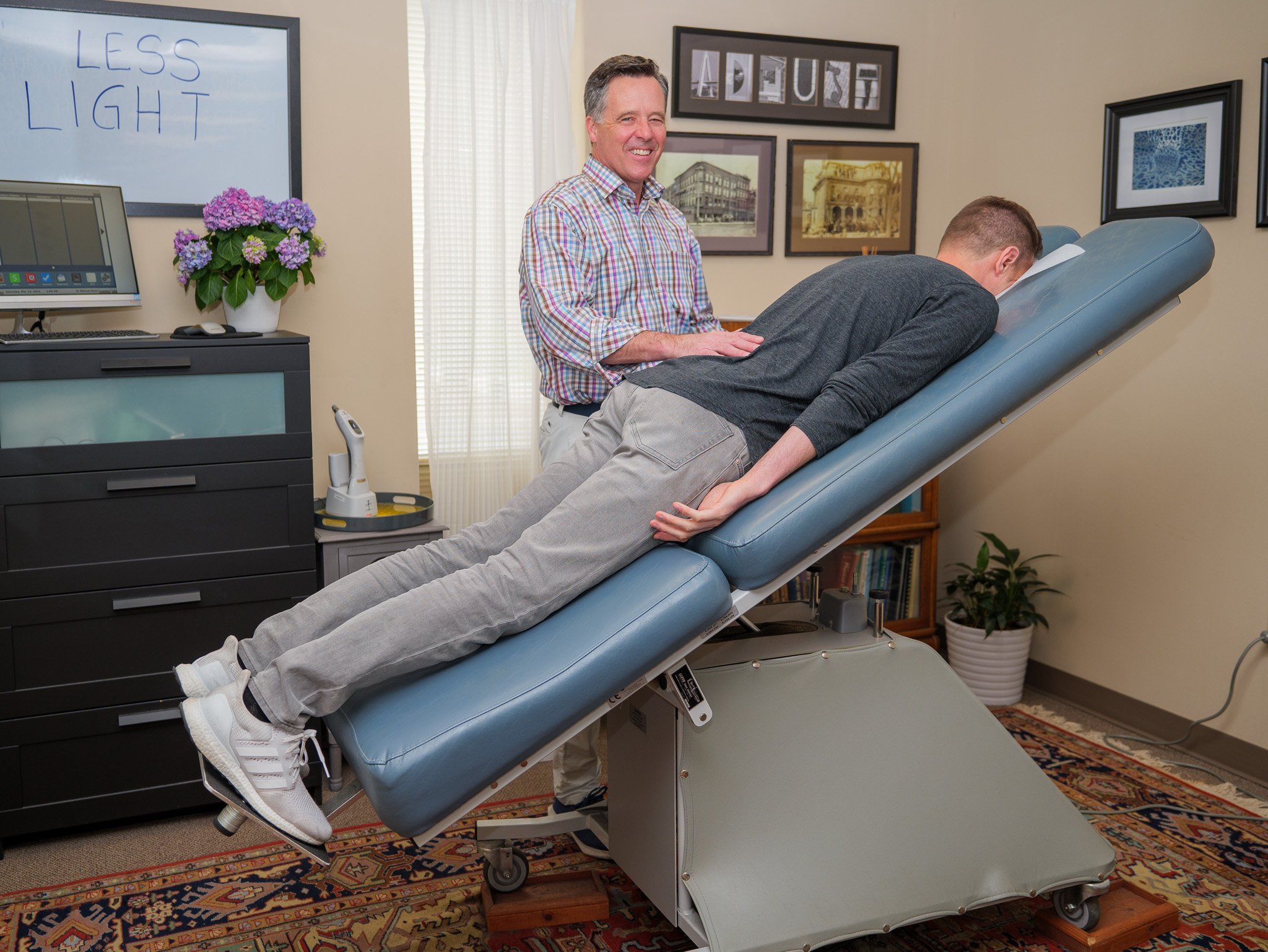 A person is lying face down on an adjustable medical examination table, while a chiropractor stands nearby smiling. The room is decorated with framed pictures and contains a cabinet with a potted plant and a whiteboard.
