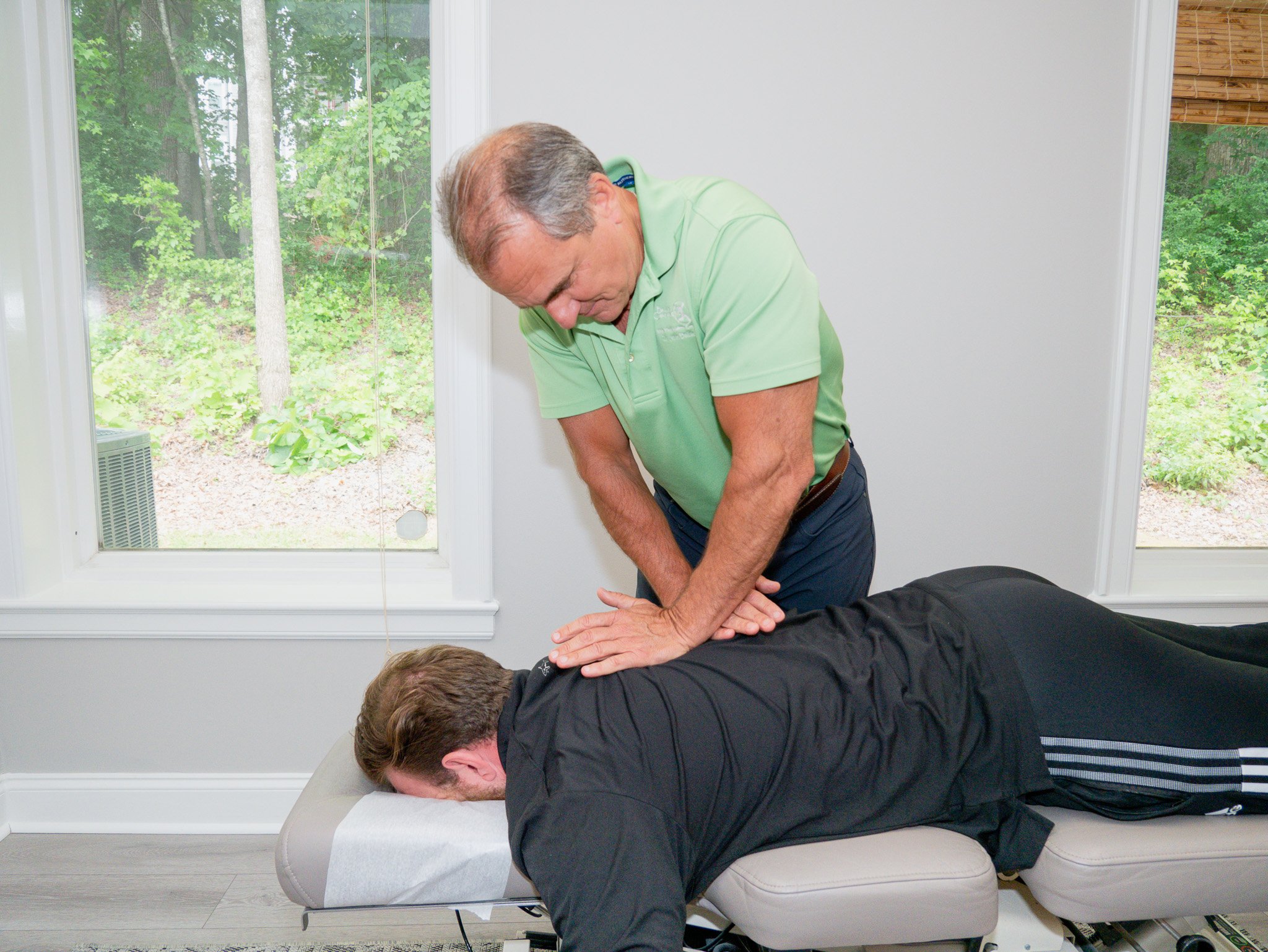 Chiropractor performing an adjustment on a patient lying face down on a treatment table in a clinic room with large windows.