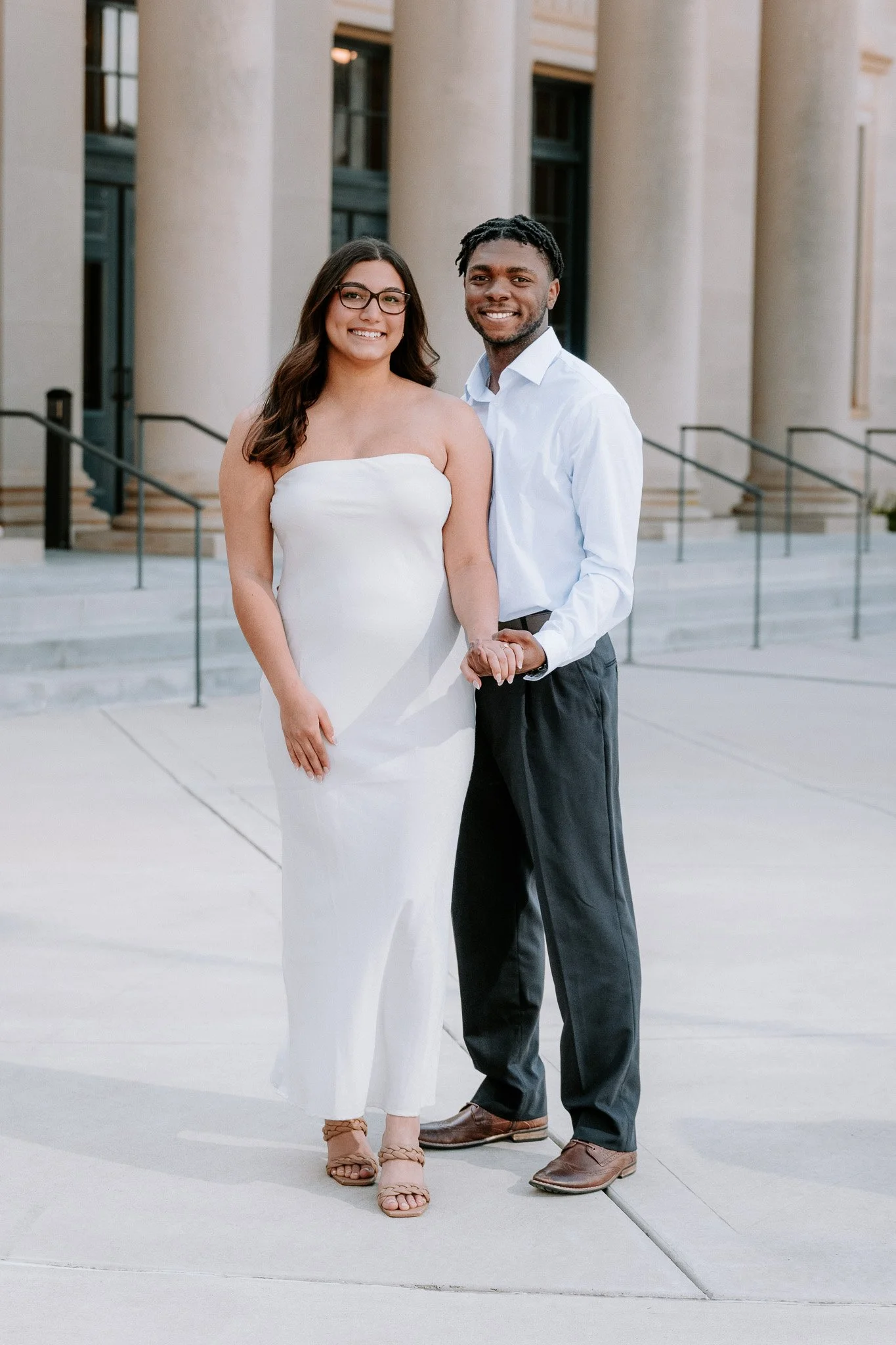 A couple stands holding hands in front of a building with large columns.  Charlotte NC Couples Engagement Portrait Photoshoot