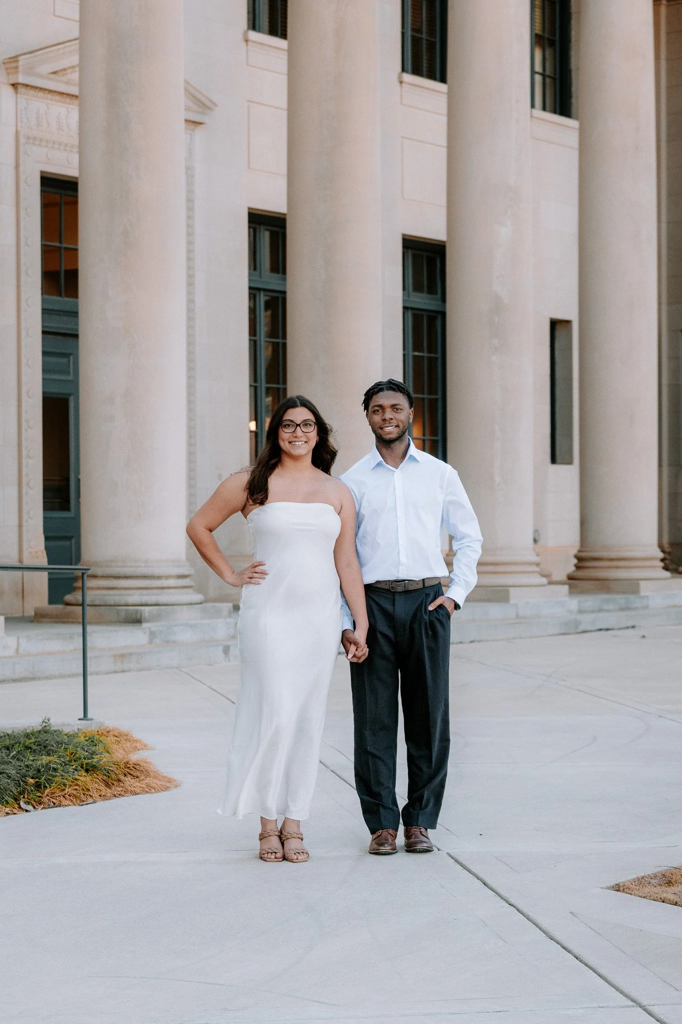 A couple standing in front of a building with columns, both dressed in formal attire. The woman is wearing a white dress, and the man is wearing a white shirt and dark pants. They are holding hands and smiling. Charlotte NC Couples Engagement Portrai