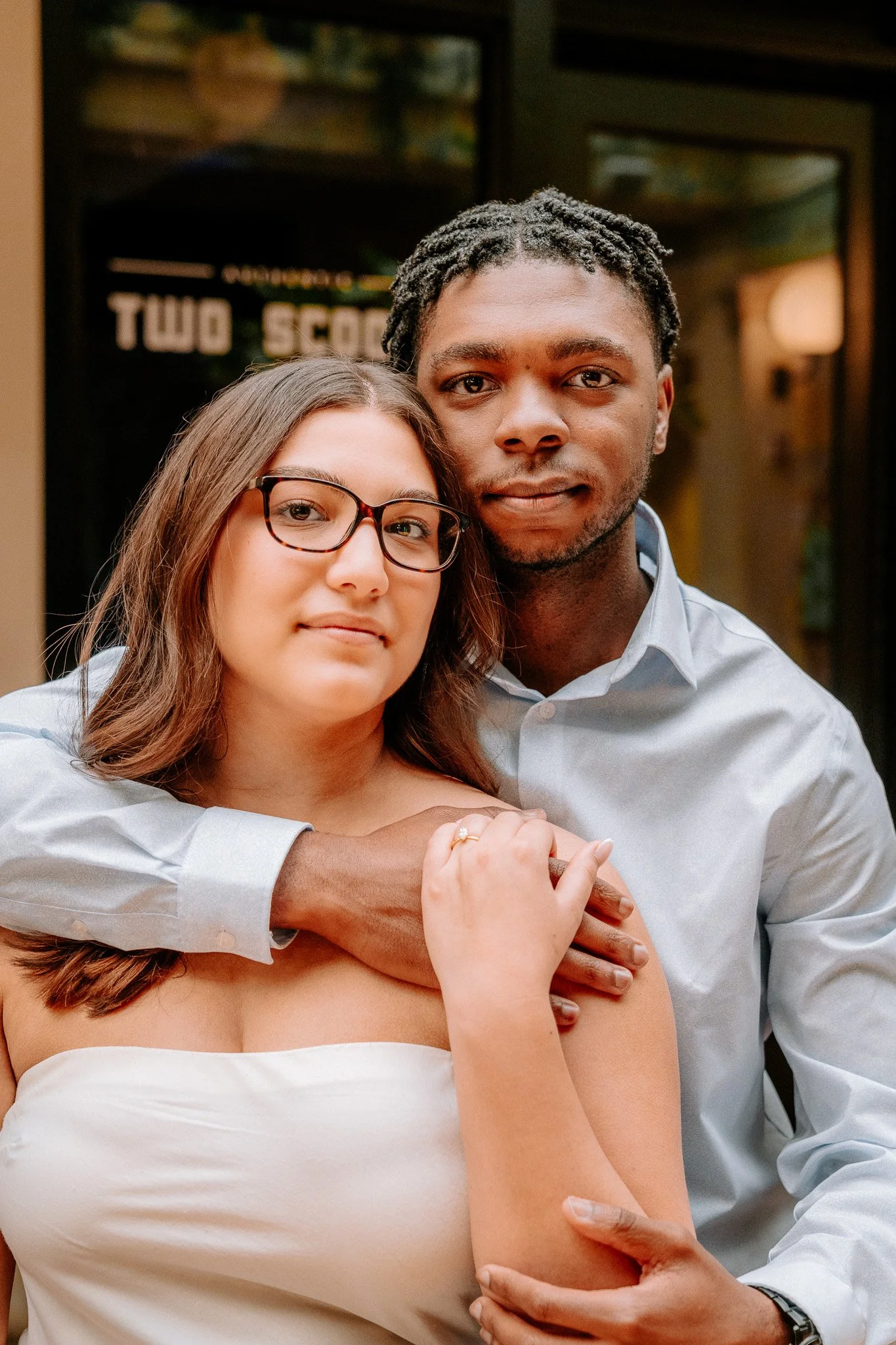A couple posing together; the woman has glasses and long hair, the man has short hair and wears a light blue shirt. They're standing in front of a dark window with text partially visible in the background.
