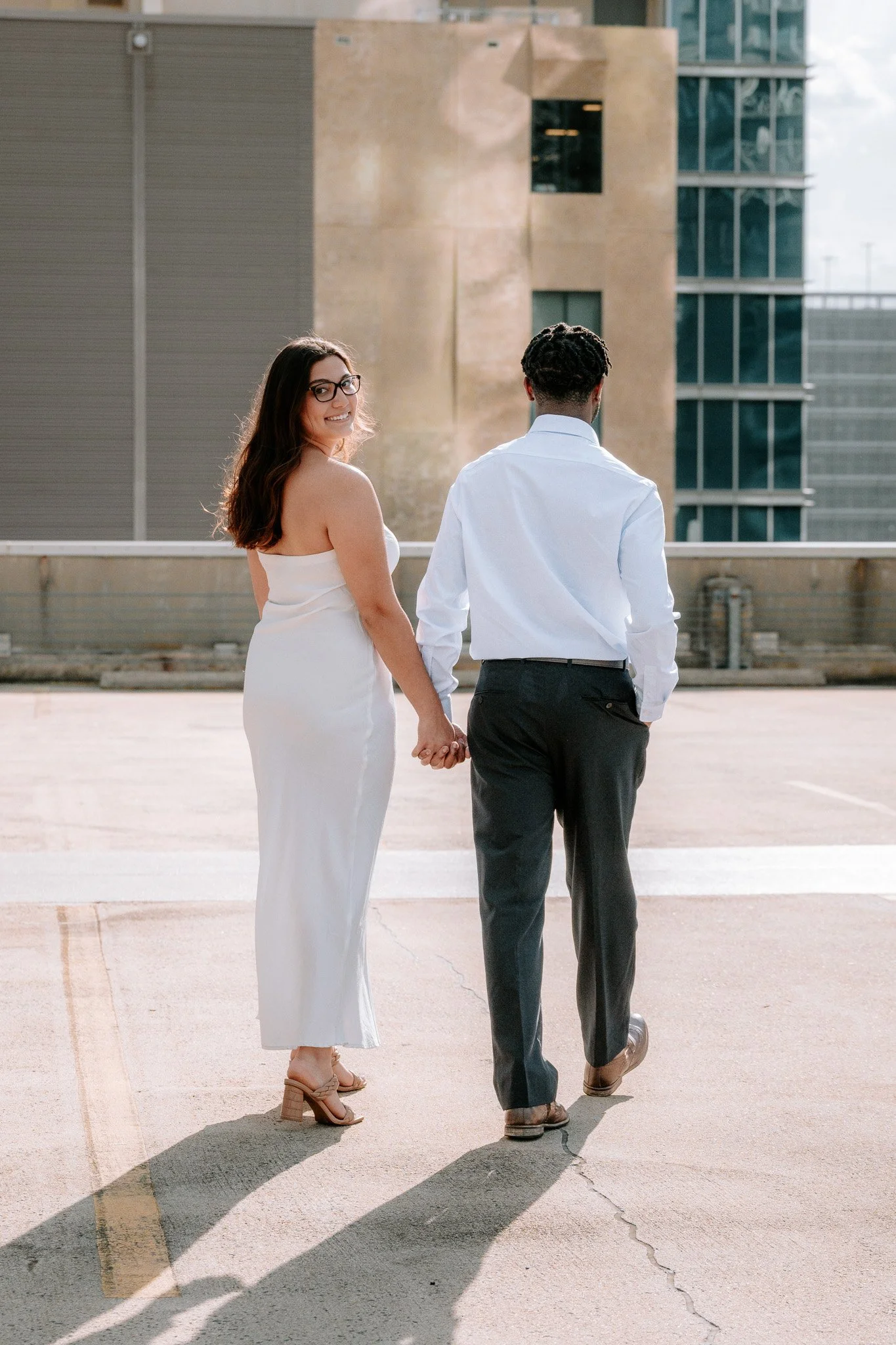 A couple holding hands, walking on a rooftop, with the woman smiling back at the camera, wearing a white dress and glasses, and the man in a white shirt and gray pants. Charlotte NC Couples Engagement Portrait Photoshoot