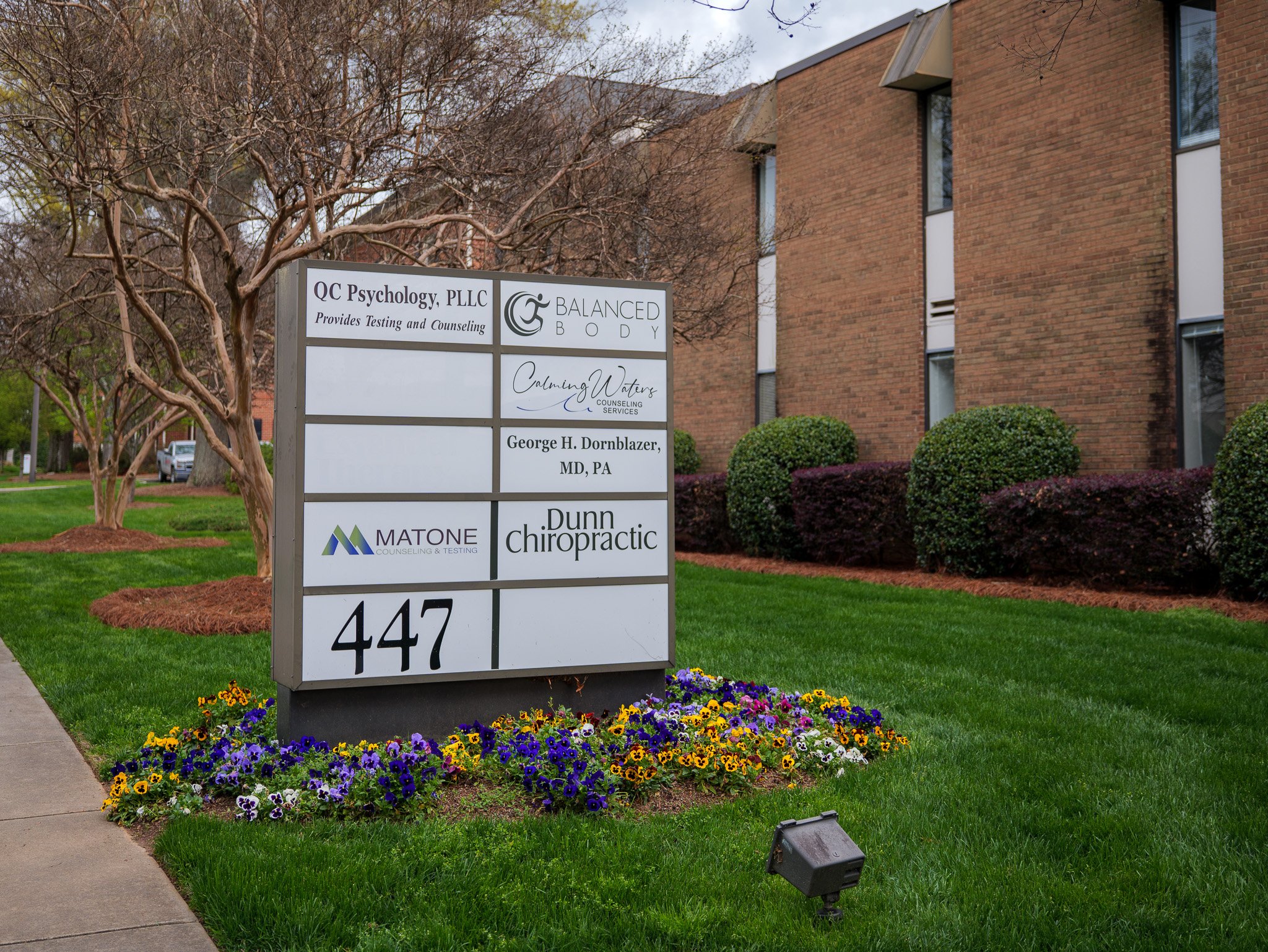 Outdoor business directory sign with multiple company names, including "QC Psychology, PLLC" and "Dunn Chiropractic," surrounded by flowers and greenery in front of a brick building.
