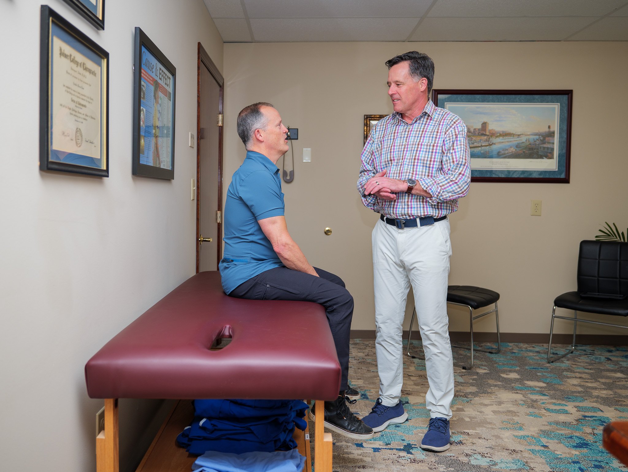 Two men in a chiropractor's office, one sitting on a treatment table, the other standing and conversing. Wall diplomas and framed art are visible in the background.