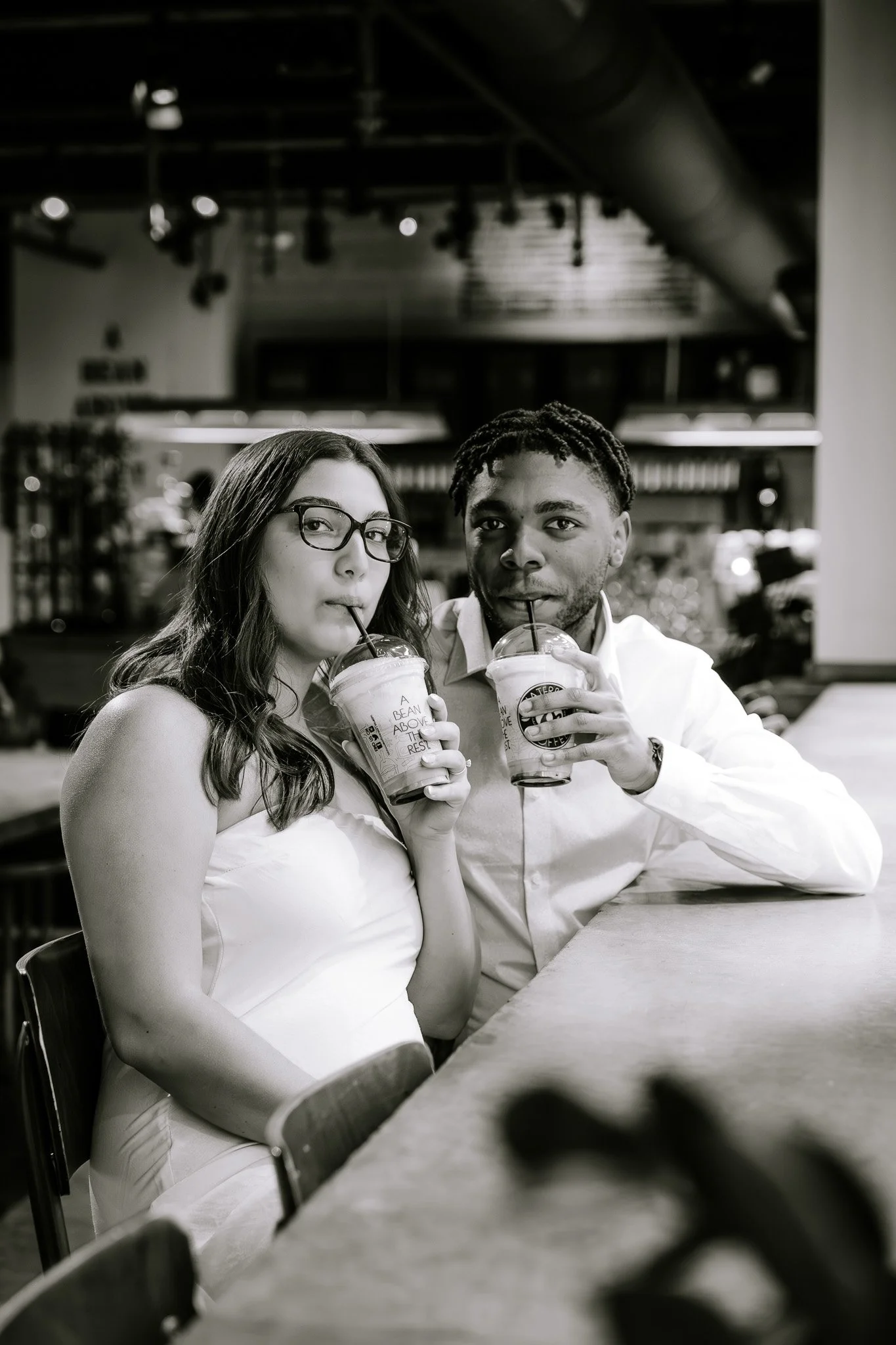 A couple sitting at a coffee shop counter, sipping iced drinks from plastic cups, black and white photo.