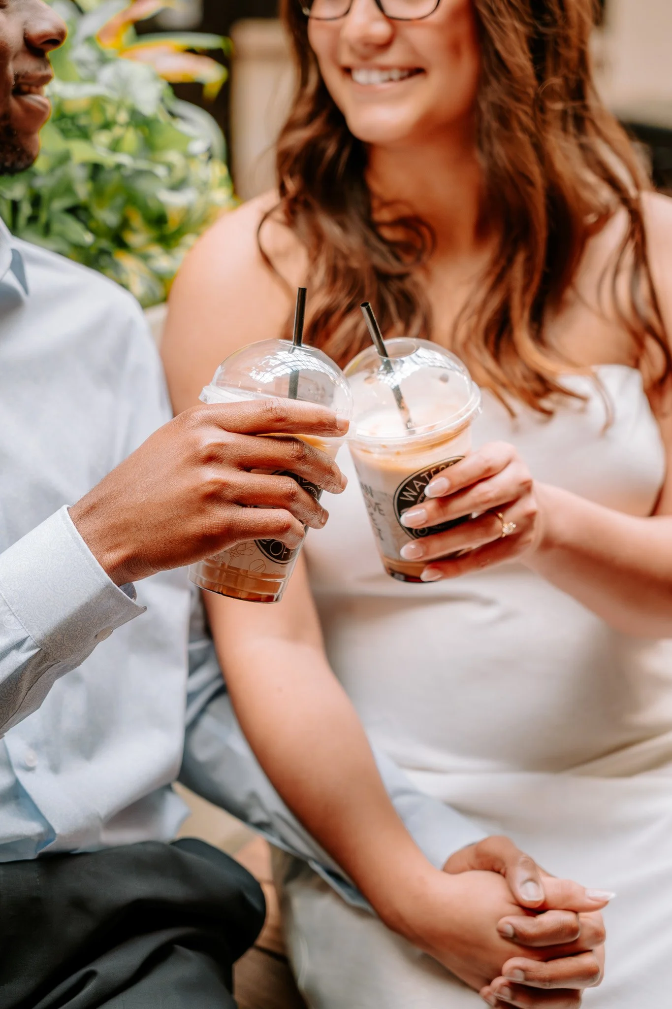Couple holding iced coffee cups in an outdoor setting, smiling and holding hands. Charlotte NC Couples Engagement Portrait Photoshoot