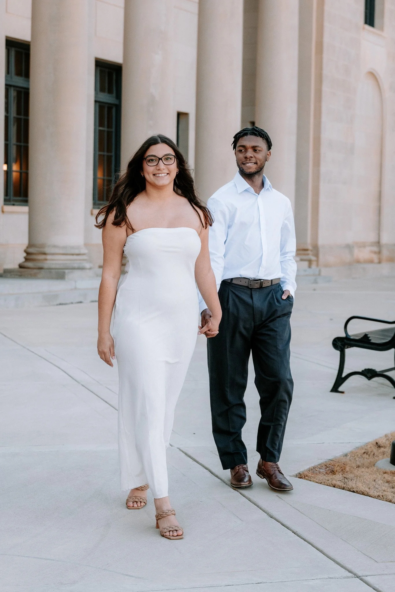 A woman in a white dress and a man in a white shirt walking outside a building with columns. Charlotte NC Couples Engagement Portrait Photoshoot