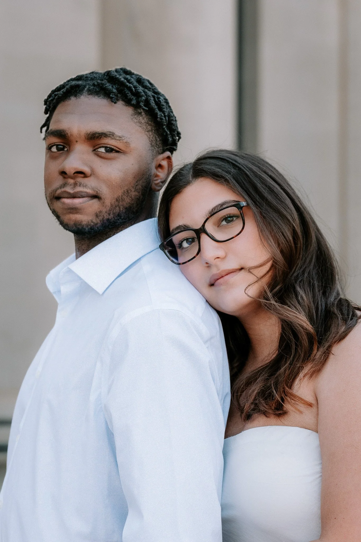 Two individuals embracing; one rests their head on the other's shoulder. Both wear white clothing against a neutral background.