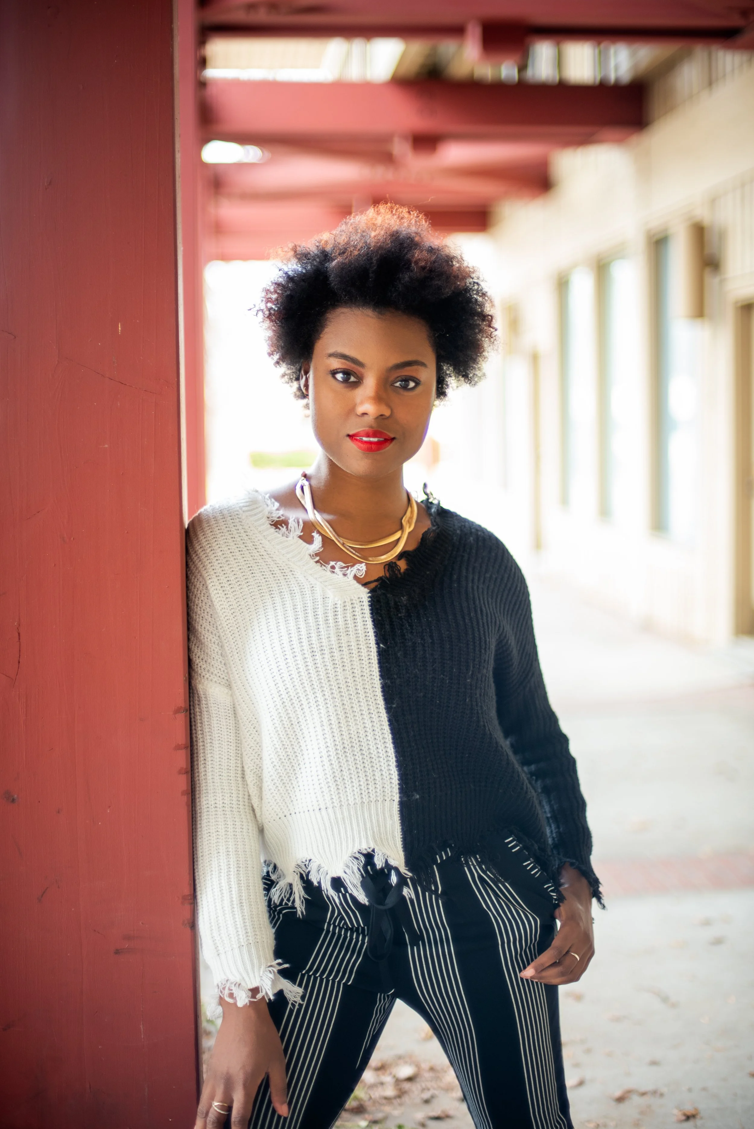 black woman with short curly hair in a black and white sweater leaning against a wooden pillar with a gold necklace and red lips